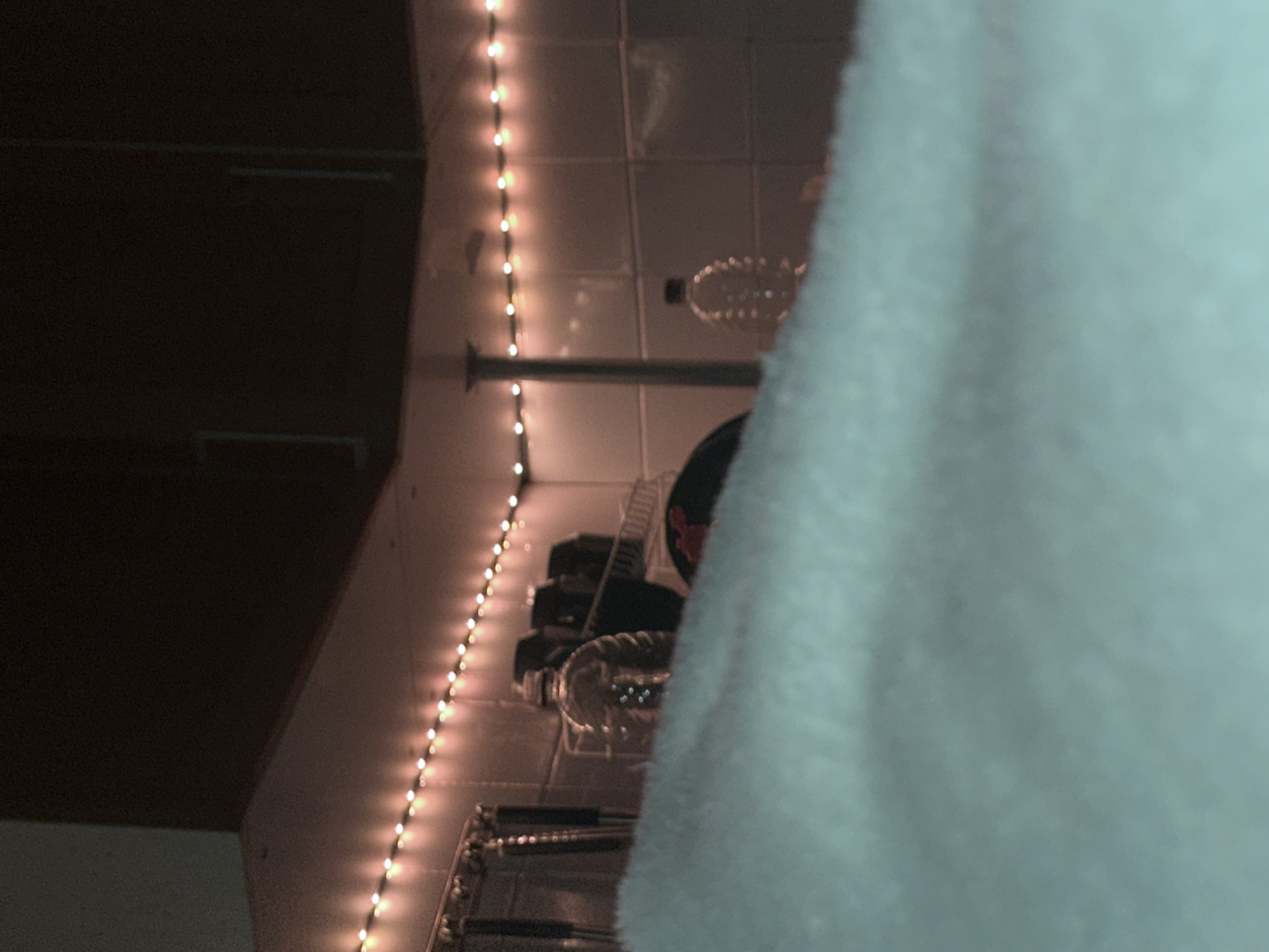 An interior scene, possibly a kitchen or utility area, is depicted in low light, attributed to the city of Moscow, Russia. The foreground is occupied by a large, blurred, light blue/cyan textile, likely a blanket or duvet, rising from the bottom edge and obscuring the lower half of the background. The background reveals a wall covered with light-colored, square tiles. A string of warm-toned LED lights illuminates the upper portion of the tiled wall, running horizontally along the underside of an upper cabinet or shelf and continuing around a visible corner. Below the LED lights, a collection of metallic kitchen utensils is visible hanging from a rail on the left. Two clear plastic water bottles, one ribbed, are present on a surface, partially obscured by the foreground textile. A dark, indistinct object with red elements is located between the bottles. A slender, vertical support structure or pipe extends from the surface to the ceiling area on the right. Dark brown upper cabinets with faint handle outlines are visible above the string lights. No individuals, specific actions, or interactions are observable.