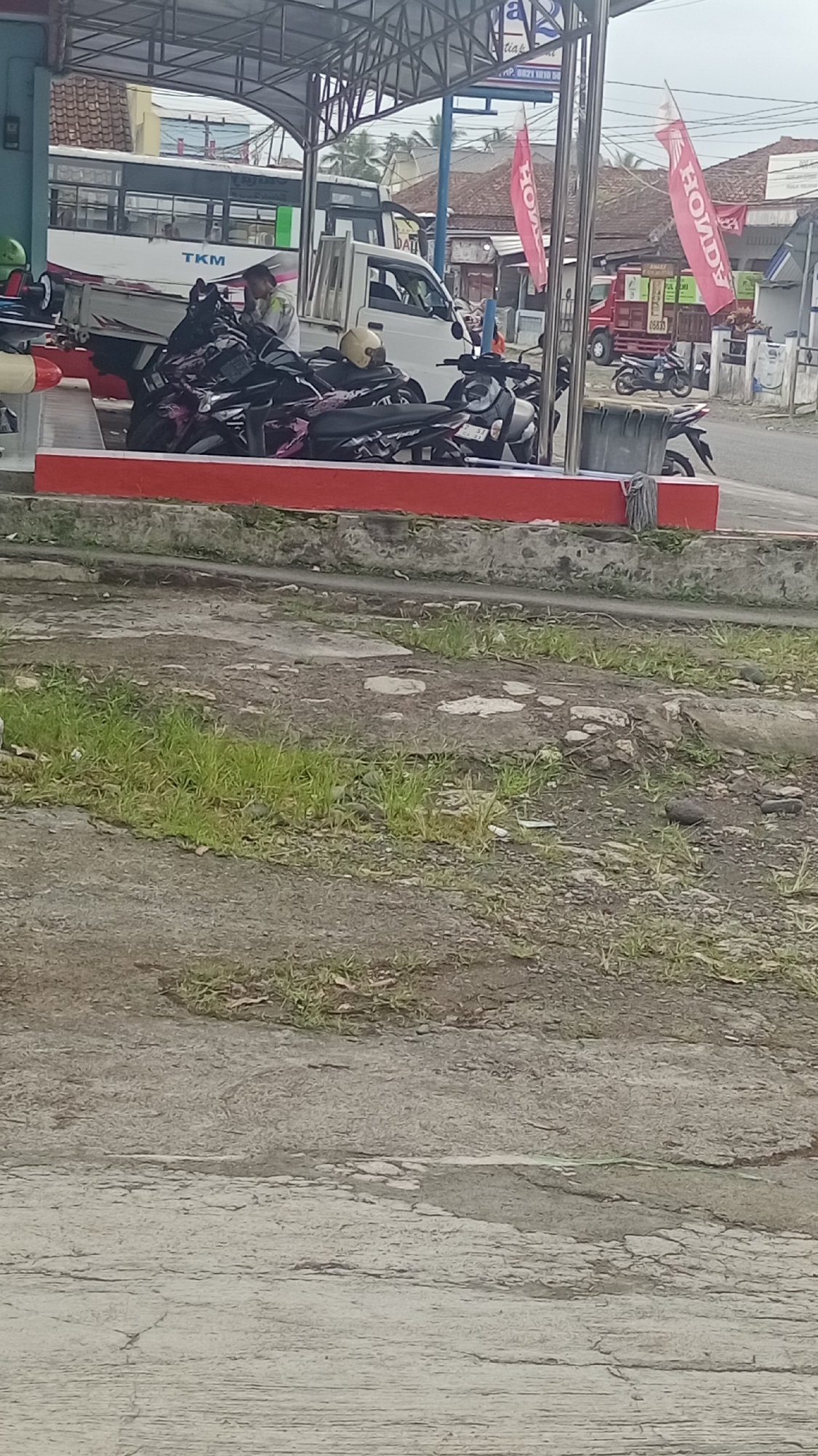 The image depicts a street scene in what appears to be Karyamandala, Indonesia. In the foreground, there is a concrete surface with patches of grass and dirt. In the midground, a red structure with metal support posts is visible, and several motorcycles are parked underneath a metal roof. One person, wearing a light-colored shirt, is seen interacting with the motorcycles. Behind this structure, a white truck with a flatbed is parked, and further in the background, a bus with "TKM" written on its side is visible. Red flags with white lettering are also present, possibly indicating a business or event. The environment appears to be an outdoor commercial area, possibly a gas station or a vehicle service area, with buildings and trees in the distance under an overcast sky.