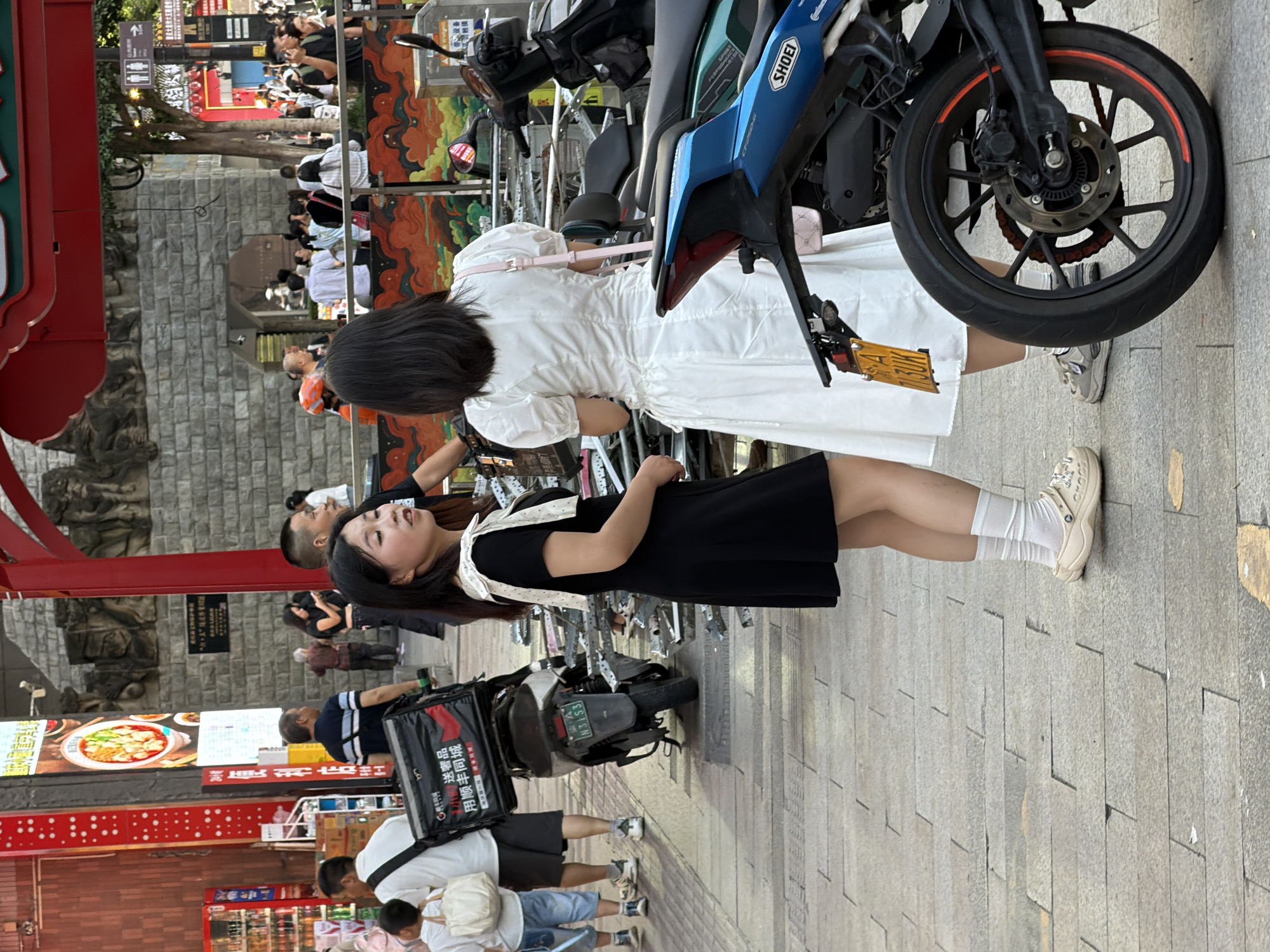 In Yuzhong District, China, two young women are standing on a paved street. The woman on the left, facing right, wears a black dress with white trim on the collar and white socks with beige clogs. She is looking to her right. The woman on the right, facing left, wears a white dress and a pink crossbody bag. She is looking towards the woman in black. Behind them, a motorcycle with a black delivery box is parked. In the background, other people, buildings, and street signs are visible. The scene appears to be a busy urban environment.