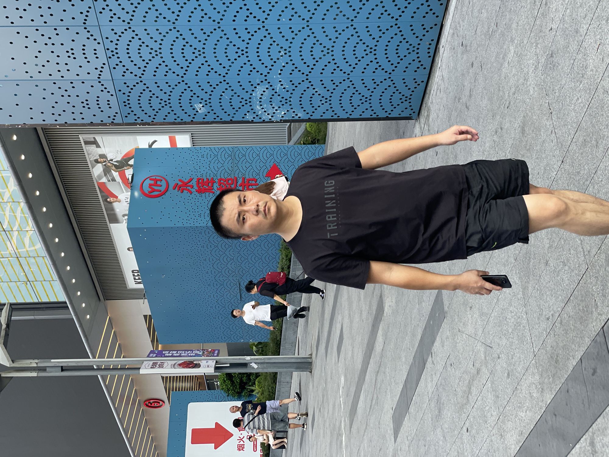 A middle-aged East Asian man wearing a black t-shirt with "TRAINING" printed on it and black shorts walks towards the camera on a paved city sidewalk. He holds a black smartphone in his right hand. In the background, several other people are visible. To the left, a woman in a light-colored dress sits on a bench. Two men in t-shirts and shorts are walking away from the camera. Further back, a man in a white t-shirt and black pants walks with a woman in a black t-shirt and red backpack. The environment includes a modern building with blue facade decorated with a pattern of circles, and a glass-fronted building in the distance. There are signs with Chinese characters and red arrows. The location is Nan'an District, China.