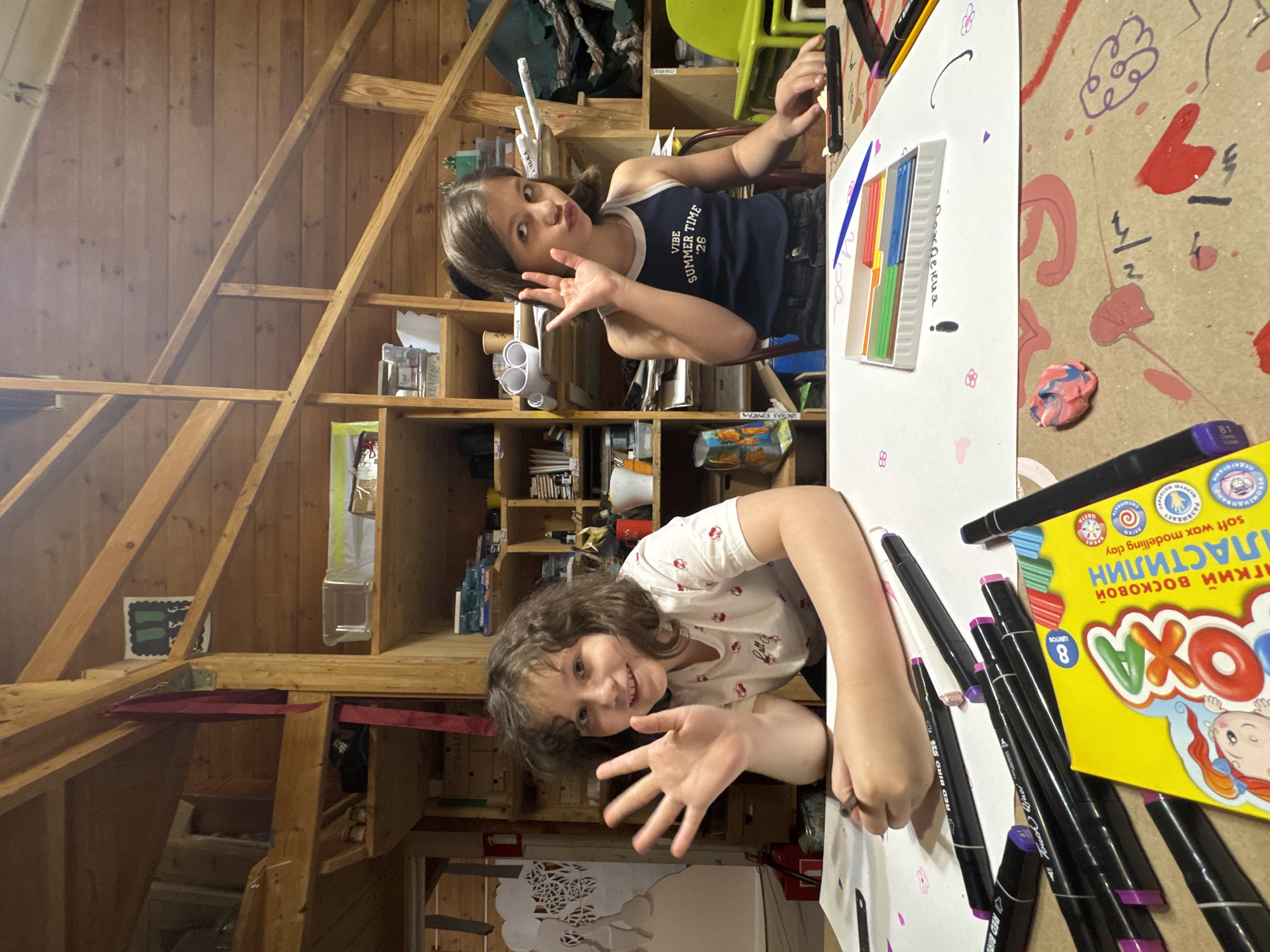 Two girls are seated at a table laden with art supplies, likely in an attic space. The younger girl, with curly hair, is at the bottom of the frame, smiling and holding out her hand. To her right, there are various markers and a box of "Okha" brand modeling clay, with text in Cyrillic script, indicating it is from a Russian-speaking region. The older girl, in a dark blue tank top with the text "Vibe Summer Time '26", is at the top of the frame, holding a marker and making a gesture with her hand. Between the girls lies a large white sheet of paper, likely used for drawing. The table surface around the paper is marked with paint and drawings. The room itself has a wooden frame and exposed beams, suggesting it is an attic or loft. Shelves along the wall are filled with miscellaneous items. The text on the modeling clay and drawing paper indicate that the location could be Moscow, Russia.
