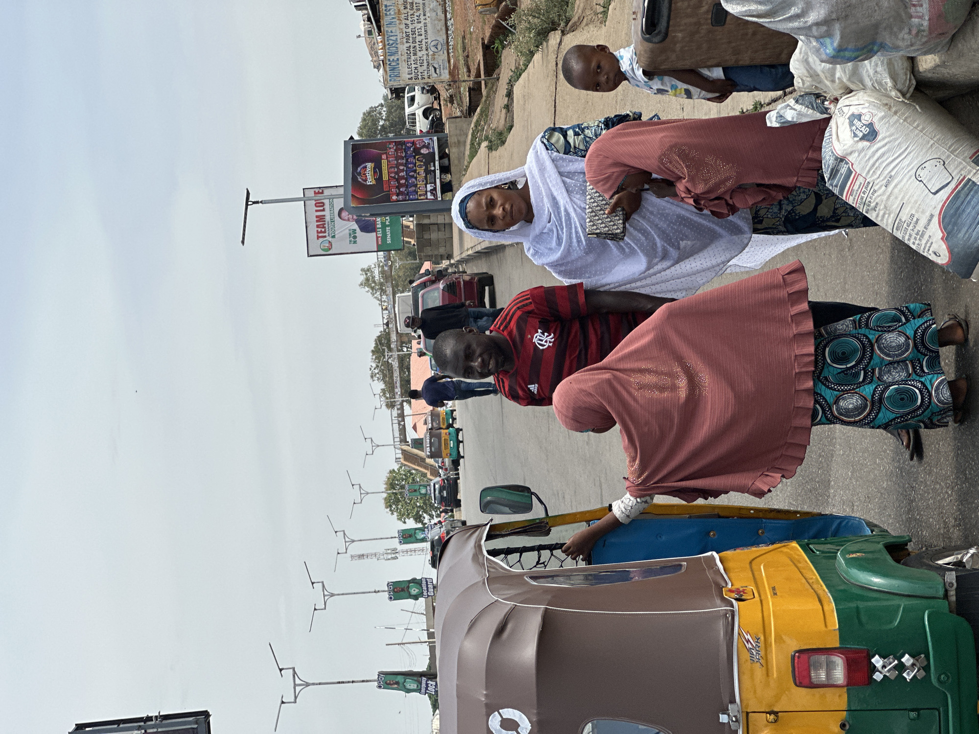 This photograph captures a bustling street scene in Jos, Nigeria, showing several people interacting near an auto-rickshaw (Keke Napep). The setting appears to be a commercial or transport hub, with paved roads, sidewalks, and buildings in the background.
In the foreground, a brown, yellow, and green Keke Napep is prominent, with a woman in a pink top and patterned skirt reaching towards its interior. Another person in a red and black striped shirt stands nearby. To the right, another woman, also in a pink top and patterned skirt, stands next to several large white sacks, one clearly labeled "BREAD FLOUR." A young boy in a patterned shirt stands behind these sacks, looking towards the camera. Further into the scene, a woman in a white headscarf and a white dotted dress looks directly at the camera, holding a small, patterned clutch bag. Behind her, a man stands near a low wall.
The activity seems to involve loading or preparing for travel, given the presence of the Keke Napep and the sacks of goods. The time of day is bright, likely midday or early afternoon, under a slightly overcast or hazy sky.
Visible text on various signs and banners includes political campaign materials like "TEAM LOVE YOUTH LIBERATION" and "HON ELI BAK SENATE PLA," as well as business advertisements such as "PRINCE MOSZYE INVEST" detailing electrical parts. Opay-branded banners advertising "Emergency Lock Guard" and "Location Guard" are also seen along the street. The "BREAD FLOUR" sack also specifies "cool, dry place" and "BATCH NO."