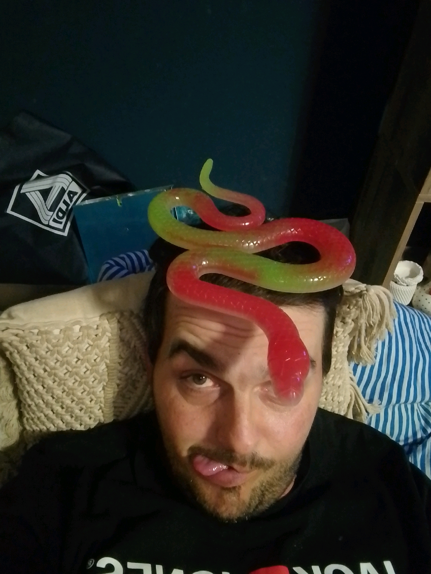 A man with dark hair and a beard is lying down with a red and green gummy snake draped over his head. His tongue is sticking out of his mouth, and one of his eyes is visible, looking directly at the camera. He is wearing a black t-shirt. Behind him, there is a beige knitted blanket and a blue and white striped cushion. To the left, a black bag with a white logo is visible. The background is dark, with some wooden shelves and a white basket on the right. This image was taken in a bedroom in Wedel, Germany.