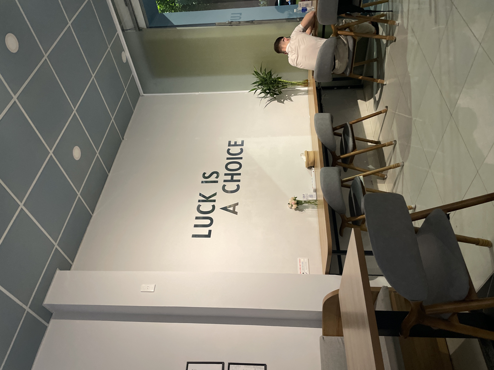 The image displays the interior of a modern cafe or similar establishment, with a clean and minimalist design. A single person is visible, seated at a long, light wooden table on the left side of the frame, wearing a light-colored shirt and appearing to look down or rest.
The right wall prominently features large, dark, sans-serif capital letters spelling out "LUCK IS A CHOICE". Below this text, on the table, a small red sign with white text is present, containing partially discernible Vietnamese characters like "QUÝ KHÁCH VUI LÒNG" (Dear Customer, Please). The table also holds small decorative plants in clear vases, one with green, bamboo-like stalks and another with delicate pink flowers. A bottle of water and a small, light-colored cylindrical container are also on the table.
The furniture includes light grey upholstered chairs with wooden frames, arranged along the table. The ceiling, at the top of the image, is a distinct blue-grey with a white diagonal grid pattern, and features recessed circular lighting fixtures. A window or glass partition at the top right reveals a glimpse of green foliage outside. The floor is covered with light-colored tiles, also arranged in a diagonal grid pattern.
The scene appears calm and quiet, likely during the daytime, with soft indoor lighting. The setting suggests a space conducive to relaxation or focused work in Bắc Nha Trang, Vietnam.