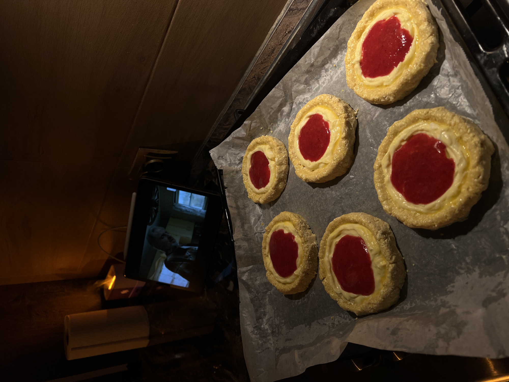 This is a forensic, matter-of-fact description of the image. The image depicts a close-up view of a baking sheet lined with parchment paper, on which six small, round pastries are arranged. Each pastry has a golden-brown crust, a creamy filling, and is topped with a dollop of red jam. The pastries are positioned on a dark countertop, possibly in a kitchen. In the background, a tablet device is visible, displaying a scene from a movie or show featuring a person with grey hair. To the left of the tablet, a roll of paper towels stands vertically, and a small, illuminated device, possibly an air freshener or humidifier, emits a warm glow. The lighting is dim, with shadows cast across the scene, suggesting an indoor setting, likely in the evening. The overall impression is of a domestic environment where someone is preparing or has just prepared baked goods while engaging with entertainment. There is no specific information in the image that indicates the location is the city of Ternopil, Ukraine.