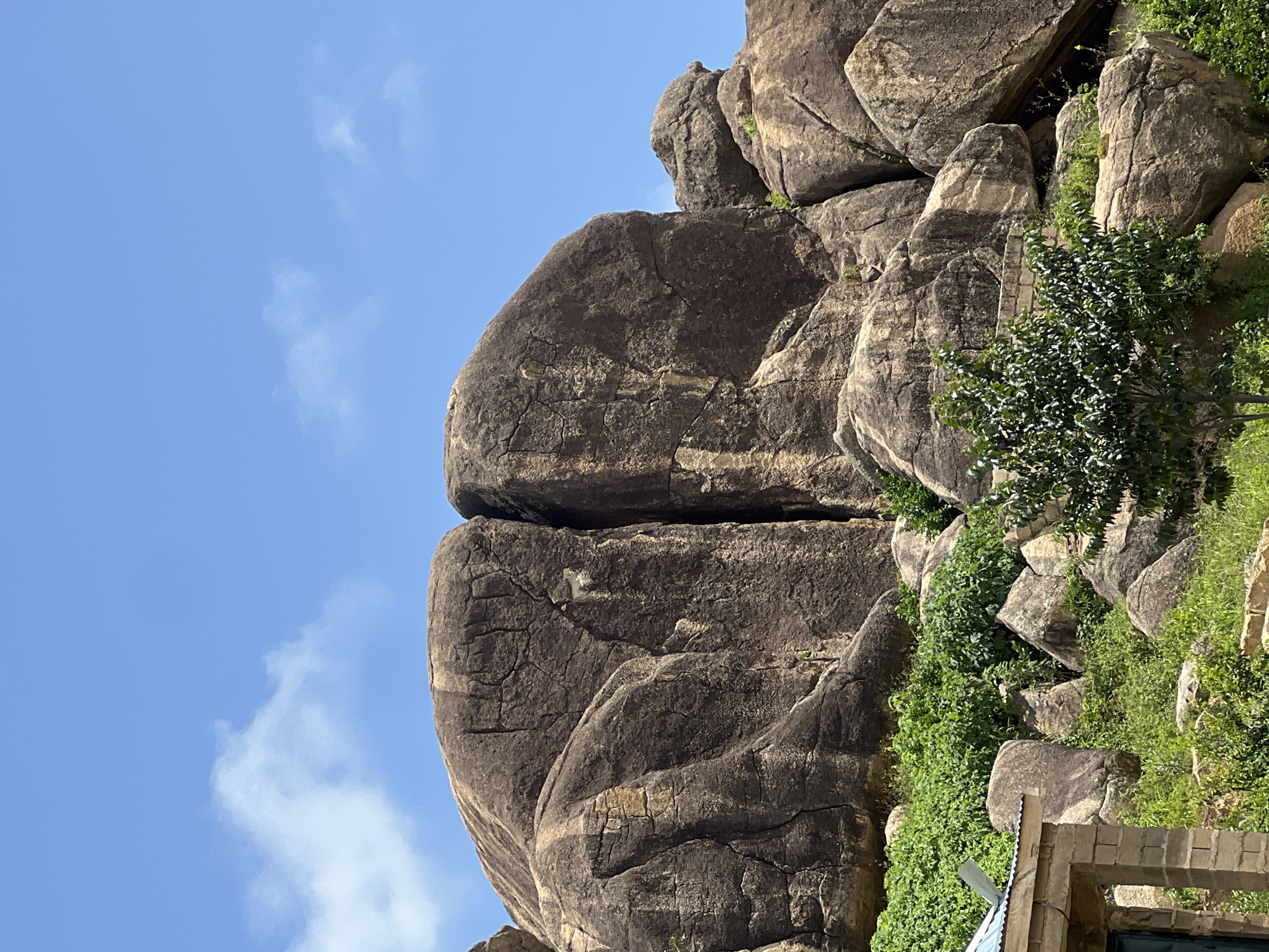 The image depicts a view of large, weathered granite rock formations that are characteristic of the landscape around Jos, Nigeria. The rocks are predominantly brown and gray, with deep crevices and a rough texture. A prominent feature is a massive rock split down the middle, creating a dramatic vertical fissure. In the foreground, there is lush green vegetation, including bushes and a small tree with dark green leaves. To the left, a portion of a simple, brick building with a corrugated metal roof is visible. The sky above is a clear, vibrant blue with a few wispy white clouds. The scene appears to be an outdoor environment, likely a natural or semi-natural area influenced by human settlement. The lighting suggests it is daytime.