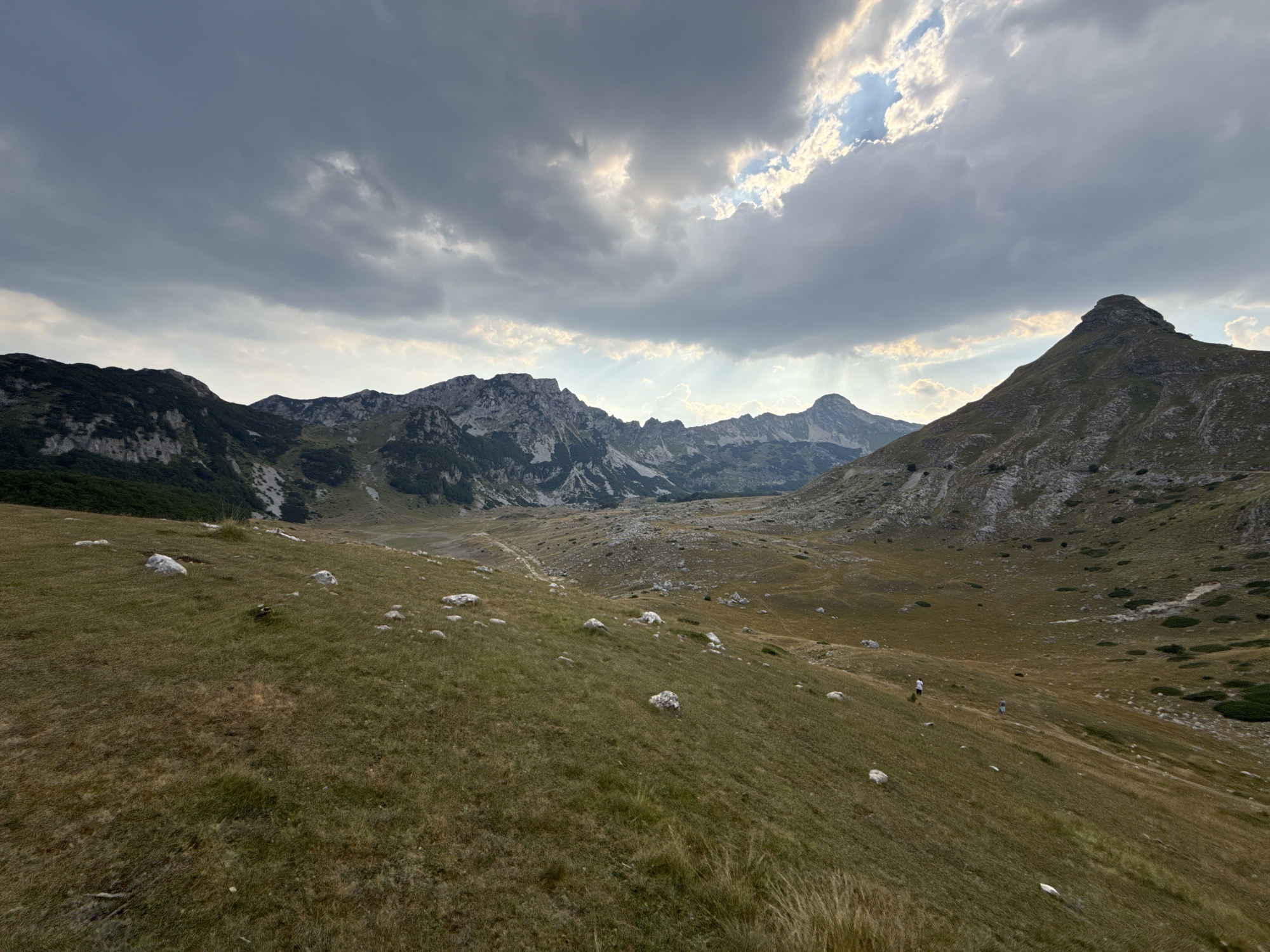 The image depicts a panoramic mountain landscape under a dramatic sky. Dark, heavy clouds dominate the upper portion of the frame, with a break revealing a sliver of bright sunlight and illuminated clouds. Below the sky, rugged mountain ranges stretch across the horizon, characterized by steep, rocky slopes and patches of dark green vegetation. The foreground is a grassy hillside dotted with white rocks of various sizes. The terrain slopes downwards from the left to the right, leading into a wide, rocky valley. Two individuals are visible on the grassy slope in the middle ground, appearing to be walking. One, in a white shirt and dark shorts, is further up the slope and to the left of the other individual, who is wearing a light blue garment and is further down the slope and to the right. The scene suggests an outdoor activity like hiking in a remote, mountainous region, possibly the area around Pašina voda, Montenegro, known for its natural beauty.