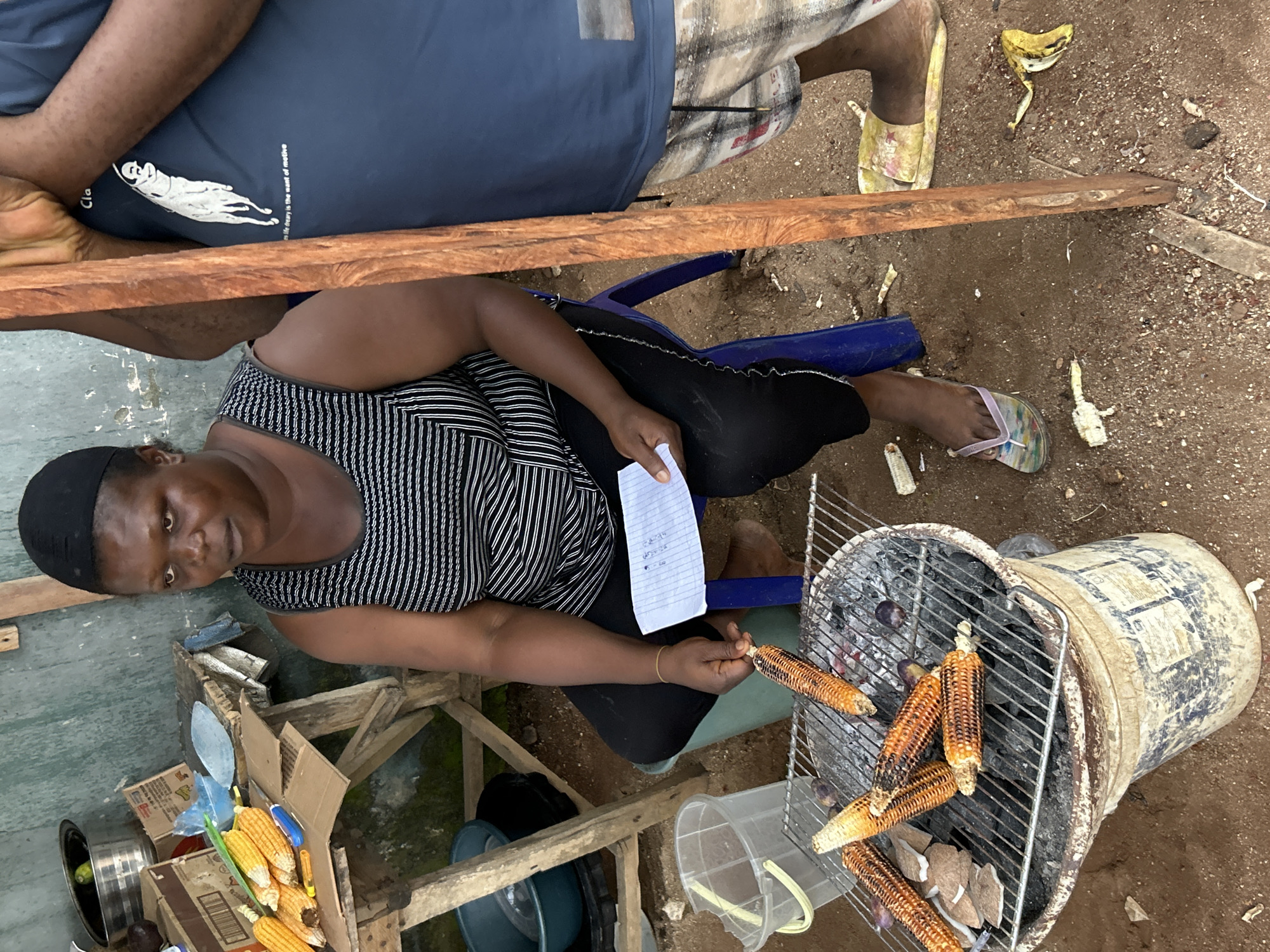 In Ihiagwa, Nigeria, an outdoor scene captures a woman tending to corn being grilled over hot coals. She is seated on a blue plastic chair, wearing a black and white striped tank top and black pants, with a black headscarf. In her right hand, she holds a piece of paper, and with her left hand, she is using a long-handled tool to adjust the corn on the grill. Several ears of corn are positioned on the metal grate of a makeshift grill, which is constructed from a bucket. The grill is filled with burning charcoal. Nearby, on a wooden table, are more ears of corn. Another person, partially visible, stands to the right, wearing a navy blue t-shirt and checkered shorts. The ground is covered in dirt and small pebbles. The environment suggests a rustic, possibly informal marketplace or roadside vendor setup.