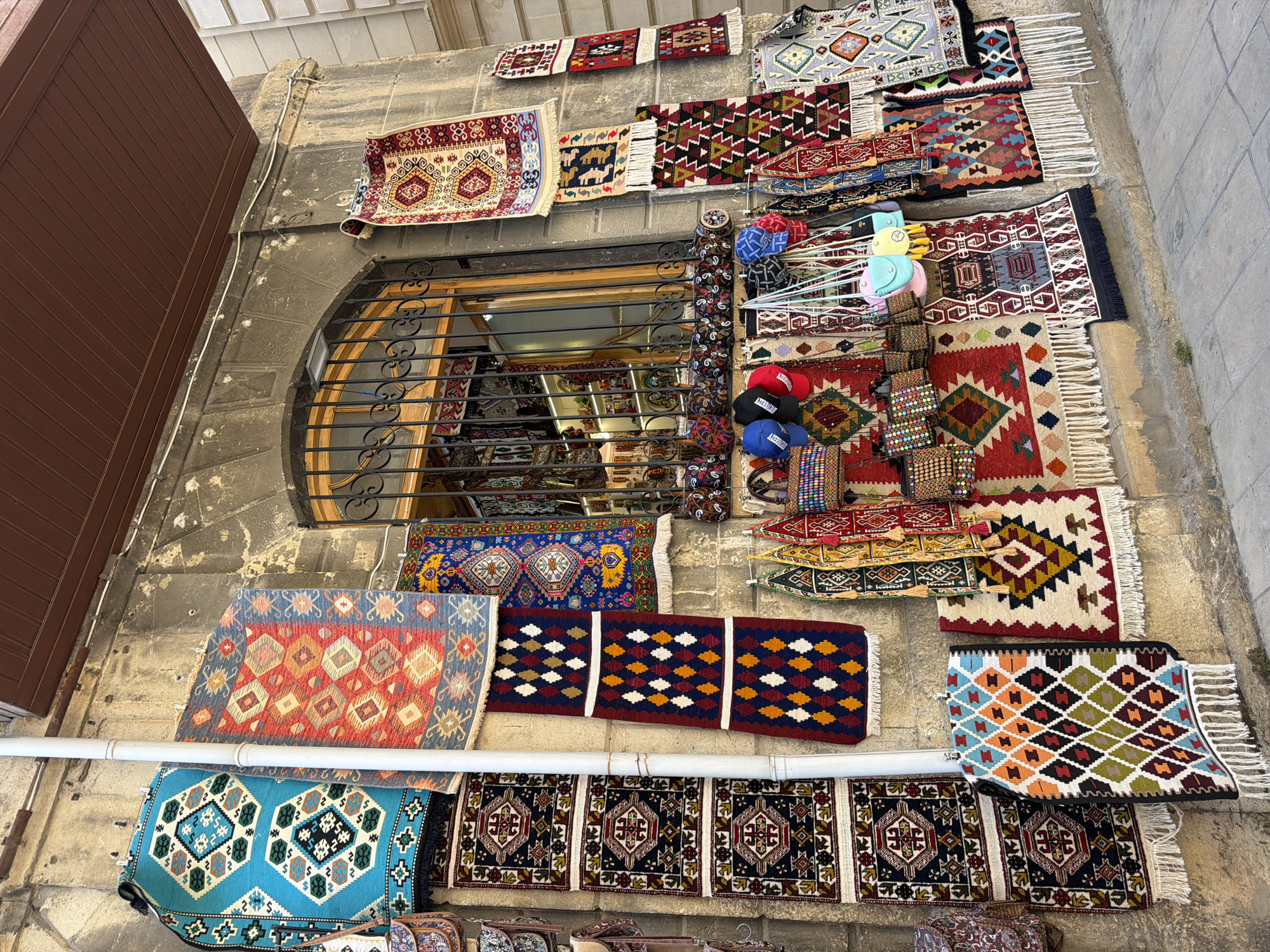 The image depicts a vibrant display of intricately patterned rugs and textile goods hanging on the exterior wall of a building in Sabail Raion, Azerbaijan. The architecture features rough-hewn stone walls and a large arched window with black metal bars, suggesting an older, possibly historic, part of the city. The textiles, predominantly in geometric patterns and rich colors like red, blue, and green, are displayed in a dense arrangement, with some hung vertically along the wall and others draped over railings. Among the rugs, there are also small bags and caps adorned with similar traditional motifs. The ground is paved with stone, and sunlight illuminates the scene, casting subtle shadows. There are no people visible in the immediate foreground, but the overall impression is of a bustling marketplace or a shop exterior showcasing local crafts.
