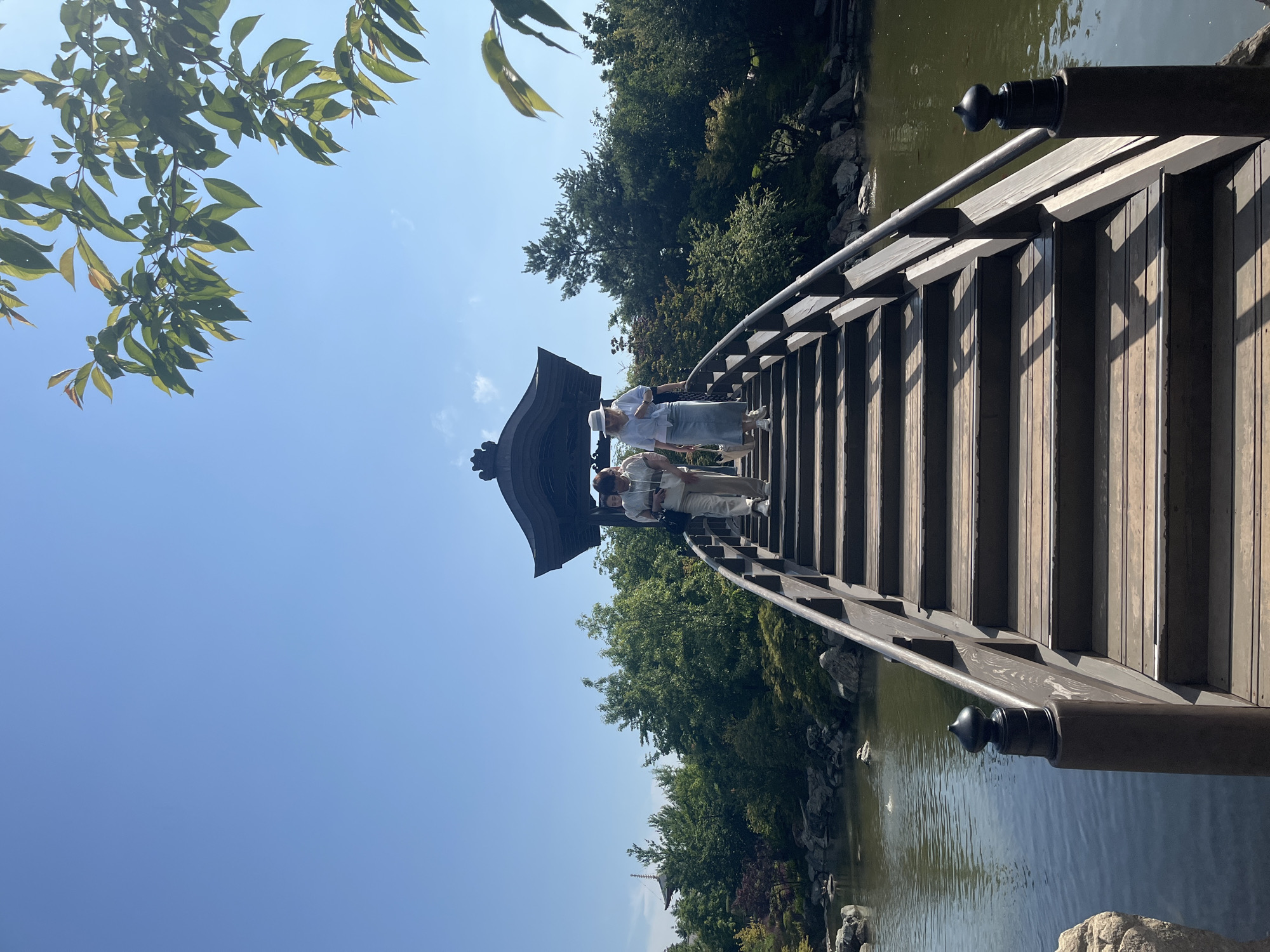 This is a vertical, eye-level, outdoor photograph of a wooden bridge arching over a pond in a Japanese garden. The sky is clear and blue, with some green leaves from a tree in the upper right corner. Three people are walking down the bridge. The person in the front is wearing a light blue shirt and a white hat, with a long denim skirt. The person behind is wearing a light gray hooded jacket and tan pants. The third person is mostly obscured. The bridge is made of dark wood planks and has dark wooden railings with decorative finials on the posts. The pond is filled with dark water, and there are rocks and greenery around its edges. In the background, there is a dense line of trees, and a multi-tiered pagoda can be seen in the distance. The garden appears to be well-maintained and peaceful. The location is not in Krasnodar, Russia.