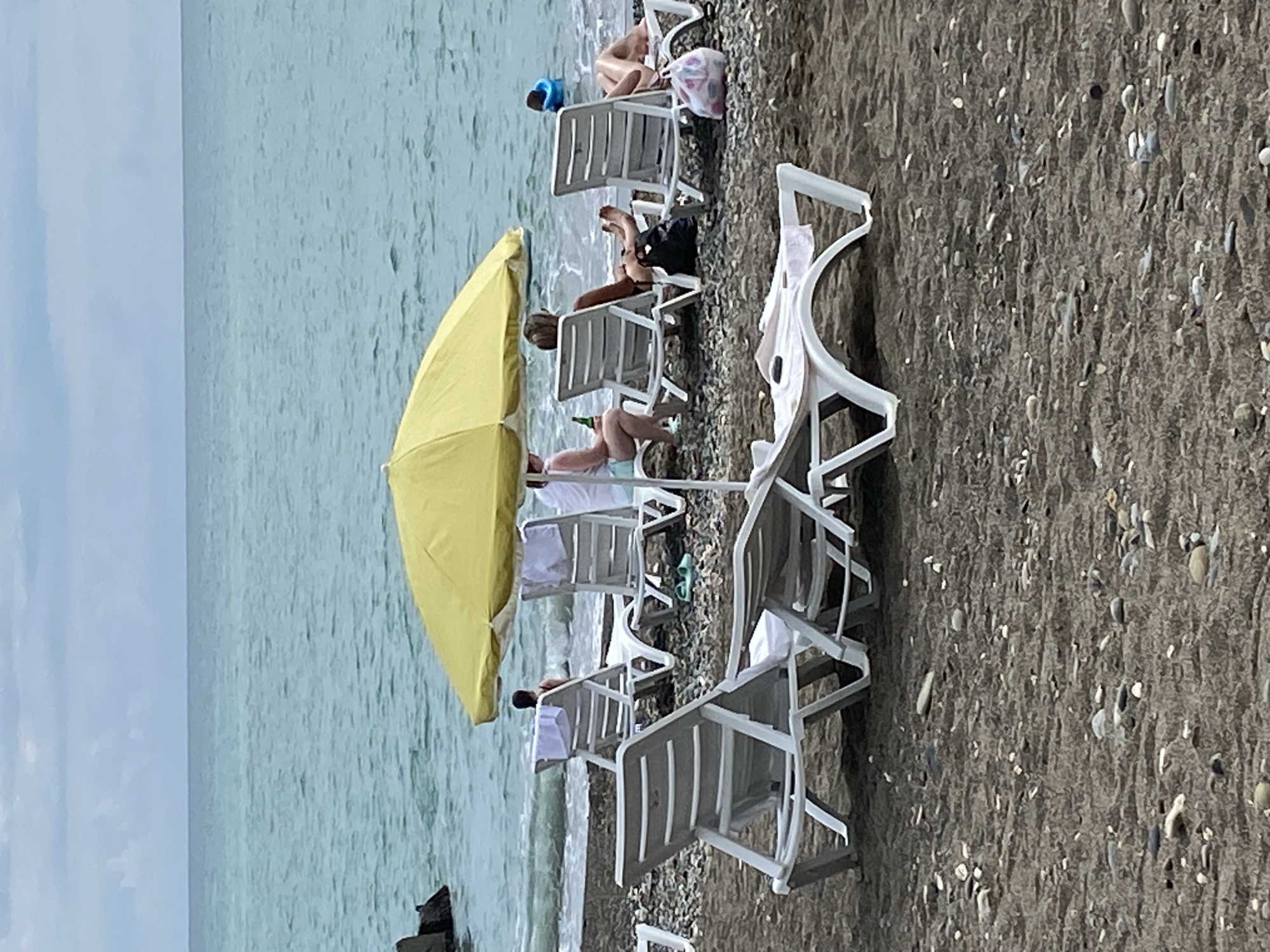 The image depicts a scene on a beach in Sokhumi, Abkhazia. In the foreground, several white plastic chaise lounges are arranged on a dark, pebbly beach. The sea, with its greenish-blue water and gentle waves, forms the background, meeting a hazy sky. A bright yellow umbrella shades a few people seated in lounge chairs further back on the beach. Some individuals are visible in the water, including one person wearing a blue floatation device. The overall atmosphere suggests a relaxed day at the seaside, with several people enjoying the beach environment.