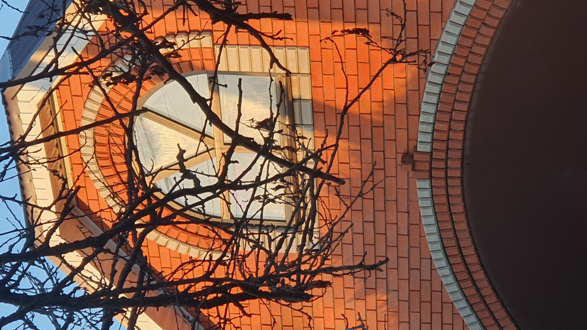The image shows the upper facade of a brick building, possibly a house, with an arched window. The brickwork is predominantly orange-red, with a decorative arch above the window featuring lighter colored bricks. The window itself is framed by a wooden structure and appears to have a frosted or textured glass. Bare tree branches with no leaves obscure parts of the facade and window. The lighting suggests it is either sunrise or sunset, casting a warm glow on the bricks. The sky above the building is a clear blue. The image is taken from a low angle, looking upwards. The location is identified as Ust-Labinsk, Russia.