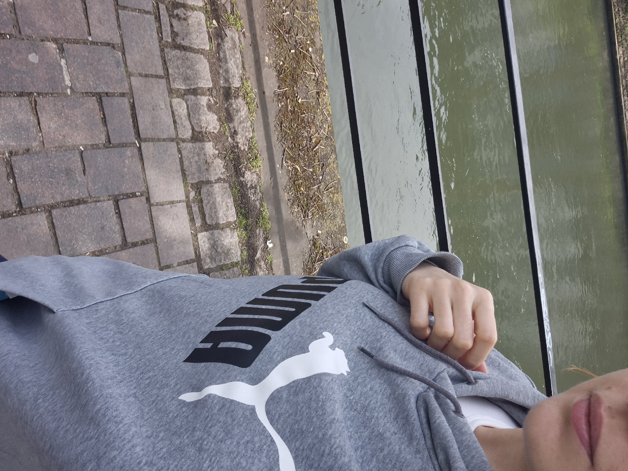 The image shows a person wearing a gray Puma hoodie standing beside a canal in Strasbourg, France. The person is leaning over a railing separating the walkway from the water. The walkway is paved with reddish-brown cobblestones. To the right of the walkway is a narrow strip of vegetation debris followed by the canal water, which is green-grey in color. Metal railings run along the canal. The person's hand is resting on the hoodie. The person's mouth is visible in the lower right corner.