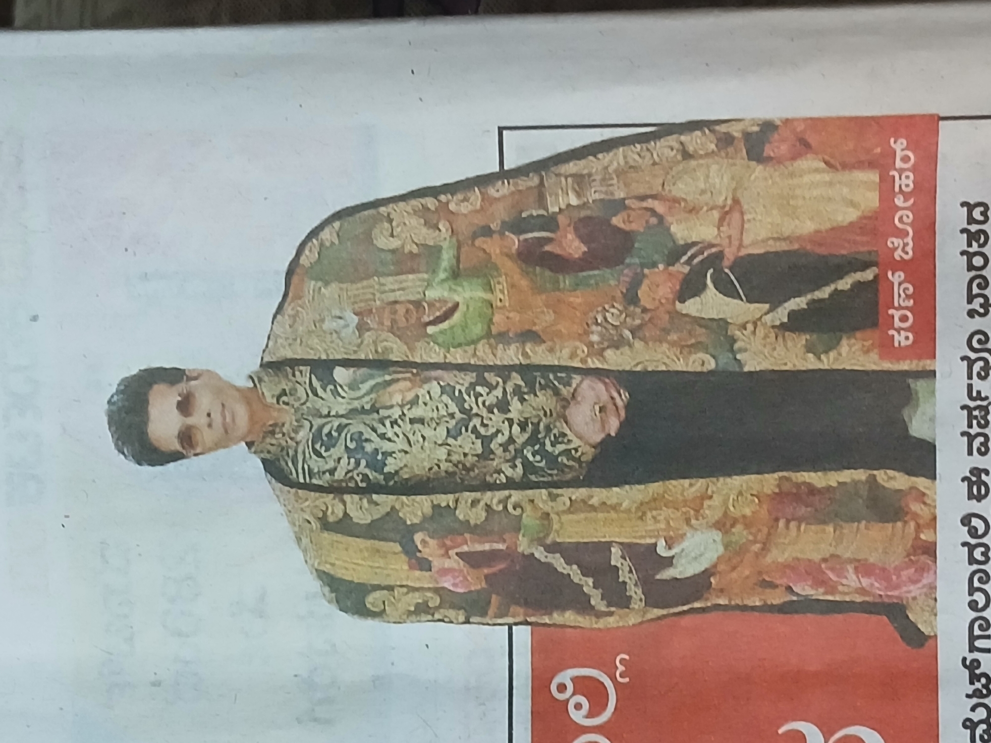 A man is photographed in what appears to be a print publication. He is wearing a heavily decorated robe or shawl. Text is visible in Kannada next to the image, reading "Karan Johar." The city is not discernible from the image.