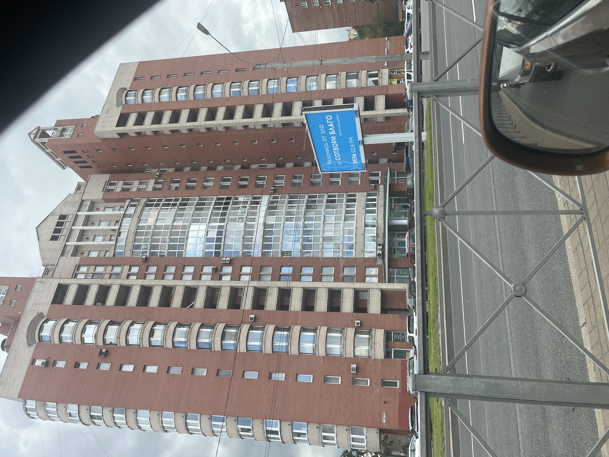 This image captures a view of a large, multi-story brick building in Saint Petersburg, Russia. The building features a mix of red brick and lighter colored facade elements, with numerous windows and balconies. A large blue billboard with Russian text and a logo is prominently displayed on a pole in front of the building. The billboard reads "УКЛОНИСЬ ОТ ЗЛА И СОТВОРИ БЛАГО" which translates to "Turn away from evil and do good." Below this, it says "ВЕРА 92.9 FM" indicating a radio station. The building appears to be a residential complex or office building. In the foreground, there is a metal fence, a sidewalk, and a road with cars. The sky is overcast. The reflection in the car's side mirror reveals more of the road and other vehicles. The overall scene suggests a typical urban environment in Saint Petersburg.