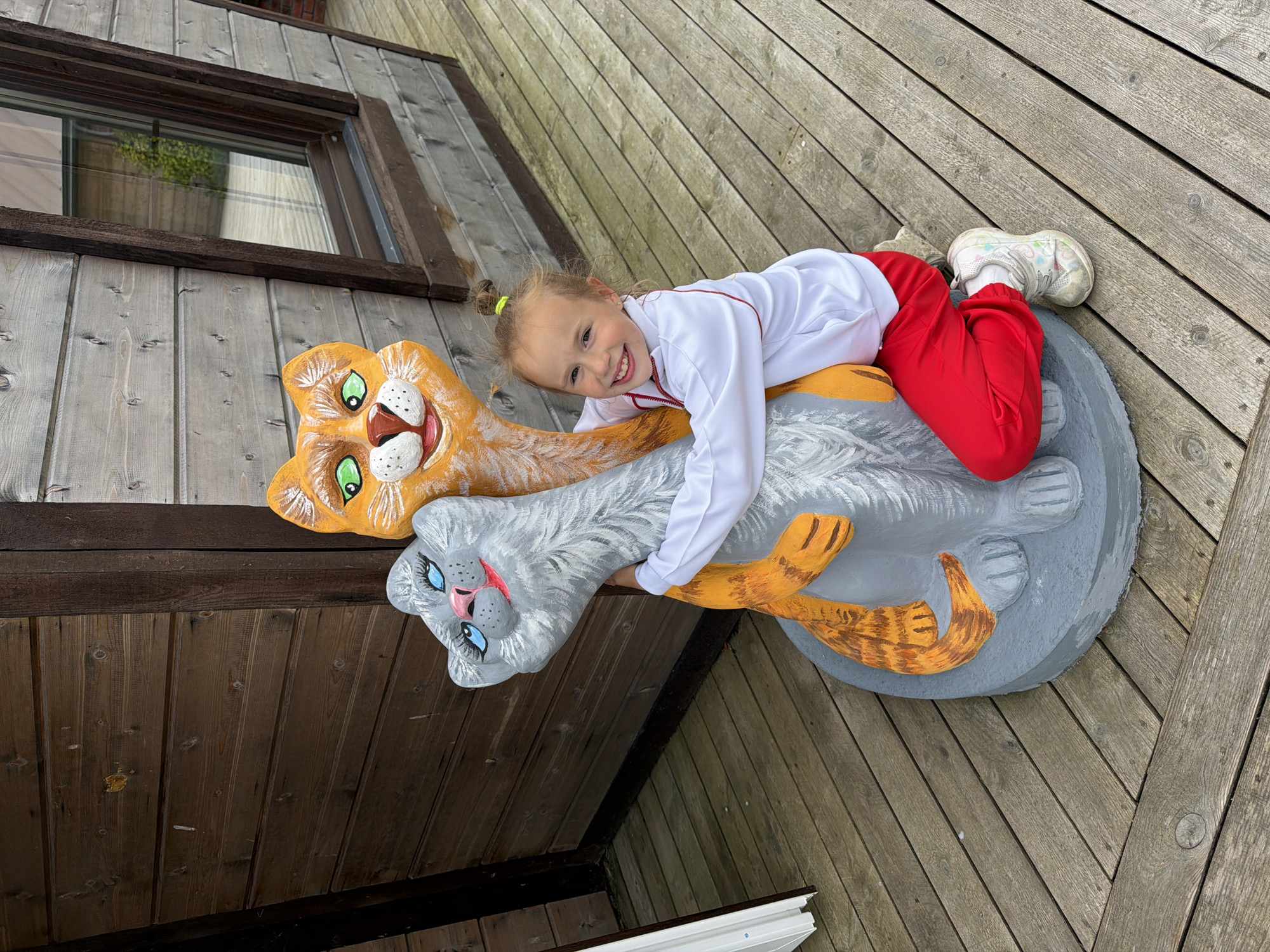 A young girl with blonde hair pulled into a ponytail, wearing a white zip-up jacket with red trim and red pants, is kneeling on a wooden deck and hugging two cartoon cat sculptures. The sculpture on the left is gray with blue eyes and a pink nose, while the sculpture on the right is orange with green eyes and a pink nose. The cats are intertwined. The background shows a wooden building with horizontal planks. The scene appears to be outdoors, with the wooden deck extending into the foreground. The overall impression is one of playful interaction between the child and the decorative sculptures. The image is taken from a slightly elevated angle, looking down at the girl and the sculptures. The lighting suggests it is daytime. The specific location in Nekrasovo, Russia, cannot be determined from the visual information provided.