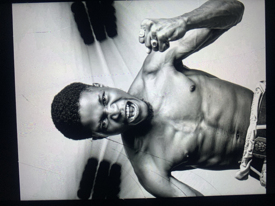 This black and white studio portrait, originating from Jos, Nigeria, features a shirtless adult male of apparent African descent, positioned from the waist up against a plain, light-toned background. He has short, curly dark hair and a sparse beard. His facial expression is intense, with his mouth wide open as if shouting or roaring, teeth visible, and eyes narrowed. His muscular physique is evident, particularly in the chest, shoulders, and arms. His left arm is bent at the elbow, with the fist clenched and held near his shoulder. A ring with a prominent dark stone is visible on the ring finger of his left hand. His right arm is also bent, with the fist clenched and positioned lower, near his waist, which is partially obscured by light-colored fabric and a dark belt with a rectangular buckle. In the background, on either side of his head, are two large, blurred, dark, elongated shapes resembling human hands, one on the left and one on the right, extending towards the center. The image itself exhibits vertical scratch marks and light reflections, suggesting it is a photograph of a physical print or a screen.