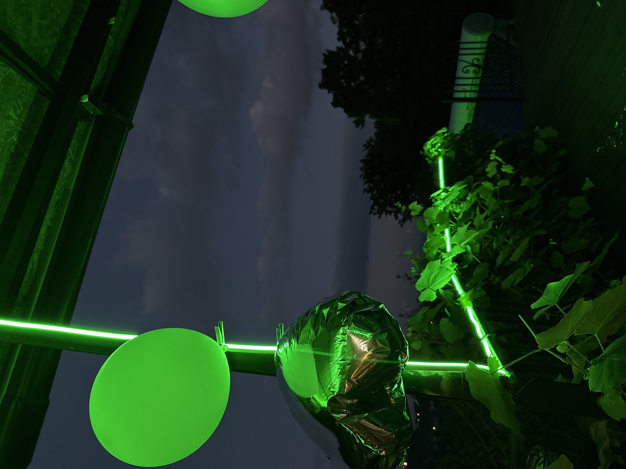 An outdoor scene at dusk or night features a green balloon and a crumpled, reflective object that appears to be another balloon, both illuminated by green LED strip lights. The lights are strung along a vertical pole and horizontally through leafy green vines. In the background, dark trees and a cloudy sky are visible, along with a white cylindrical object and a dark railing. The ground appears to be wooden decking. The location is specified as Krayevsko-Armyanskoye, Russia.