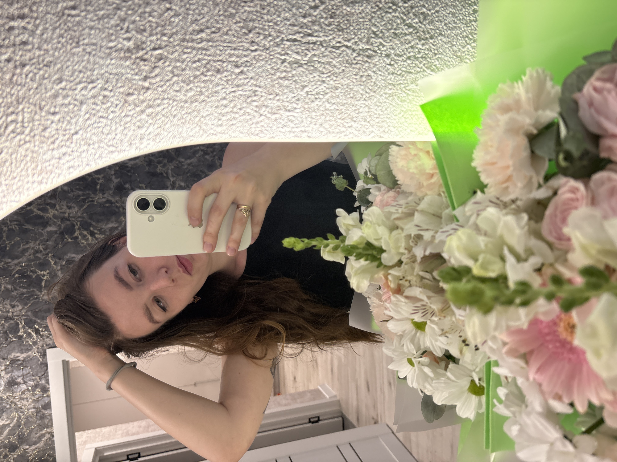 A young woman is reflected in a mirror while taking a selfie. She is holding a white smartphone with two camera lenses. She has long brown hair and is wearing a black top. Her left hand is raised, touching her hair. On her right hand, she wears a gold ring. A bouquet of white and pink flowers is positioned to the right of her reflection, partially obscuring it. The flowers are wrapped in green paper. The mirror is mounted on a black marble surface, and behind it, the floor is visible. The location could be a bathroom based on the door frame and a glimpse of the floor. There is no definitive evidence to confirm the location within the city of Izhevsk, Russia.