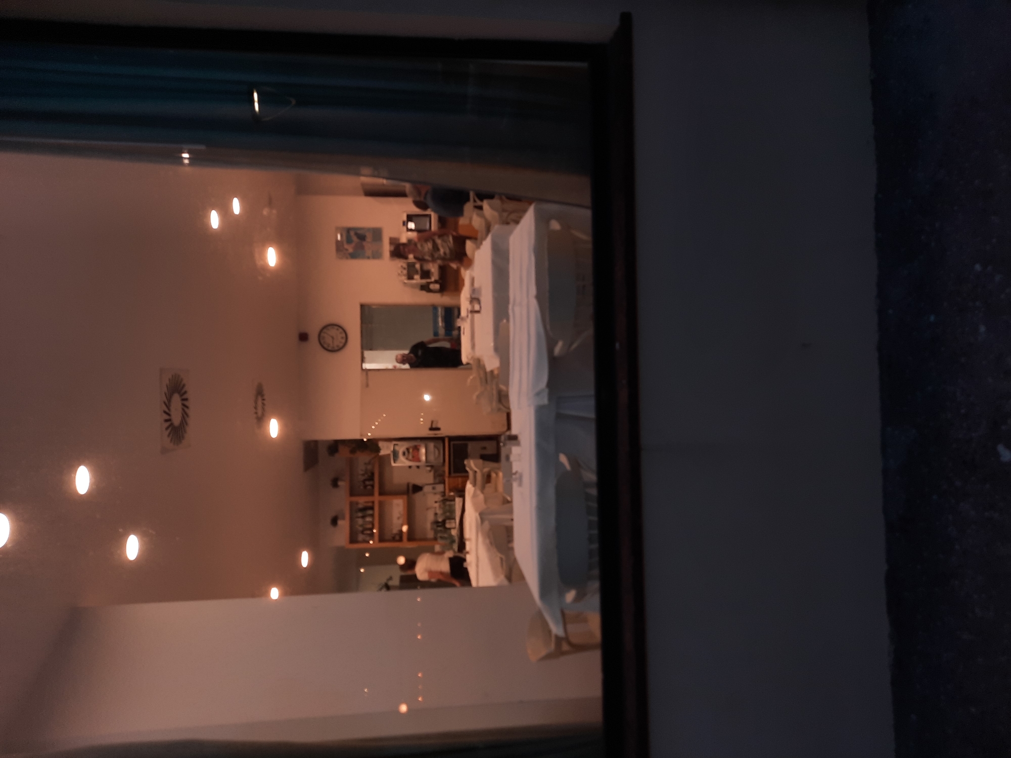 The image depicts the interior of a restaurant in Pesaro, Italy, viewed through a window. Several tables are set with white tablecloths, indicating that the restaurant is either closed or preparing for service. Lights are on in the ceiling, and there appears to be a bar area in the background with shelves stocked with bottles. Two individuals are visible inside the restaurant: one person, possibly an employee, is standing near the bar, and another person is seen through a doorway. The lighting suggests it is either early morning or evening. The overall scene is calm and orderly, suggesting a typical restaurant setting.