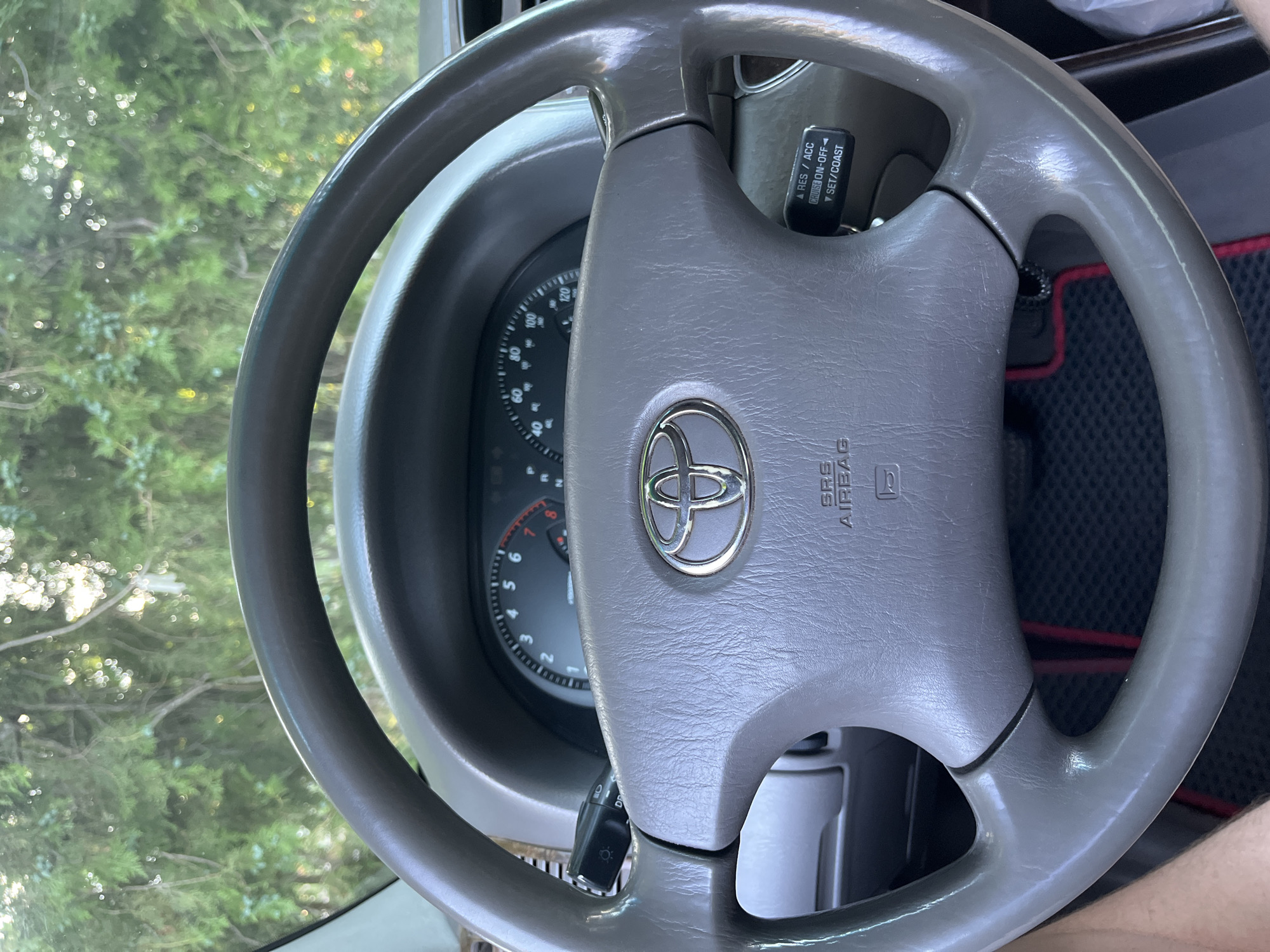 A close-up shot shows the interior of a Toyota car, focusing on the steering wheel and dashboard. The steering wheel is grey and features the Toyota logo in the center. To the left of the steering wheel, a stalk with controls for lights and indicators is visible. On the right side of the steering wheel, a cruise control panel with buttons labeled "RES/ACC," "CRUISE ON-OFF," and "SET/COAST" is present. The dashboard has a speedometer and tachometer. Through the windshield, trees and foliage are visible, suggesting the car is parked outdoors, possibly in a natural setting or on the outskirts of a populated area like Ivanivka, Ukraine. The lighting indicates it is daytime. No people are visible in the frame.
