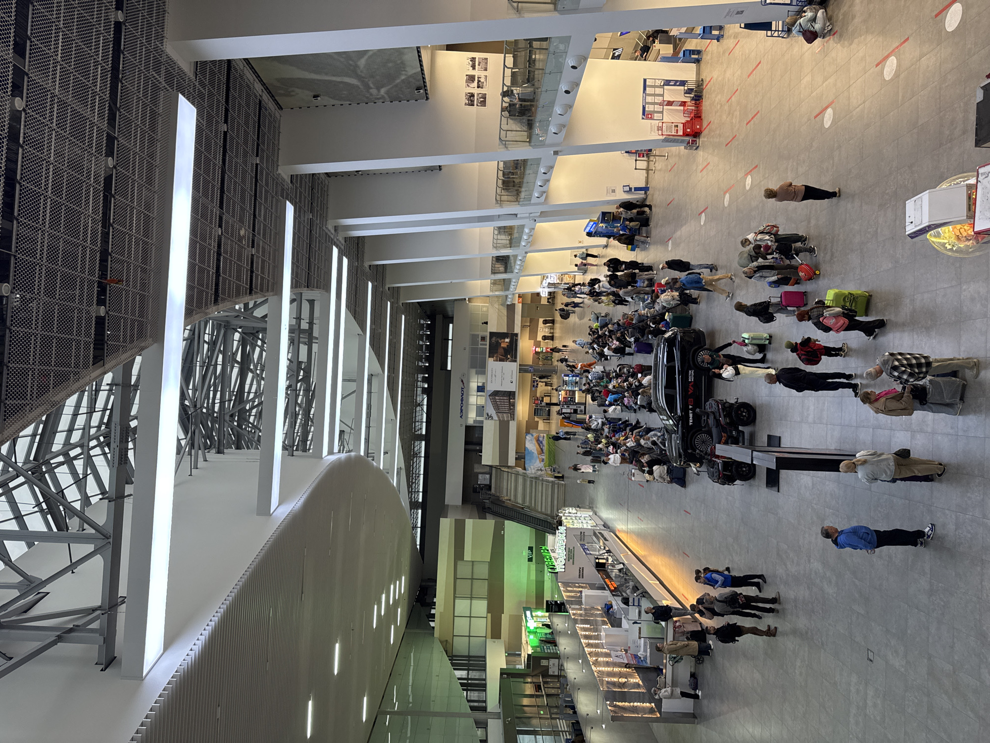 The image depicts the interior of an airport terminal. Several people are present, some queuing with luggage, others standing or walking. Lines and circles marked on the floor appear to serve as social distancing markers. The environment includes high ceilings with exposed structural elements and lighting fixtures, advertisements on the walls, and retail or service counters on one side. A black vehicle is visible amid the queuing people. Given the location is specified as Zarechnaya (Gamovskoe S/P), Russia, it can be inferred this is likely Perm International Airport, located near Gamovskoe.