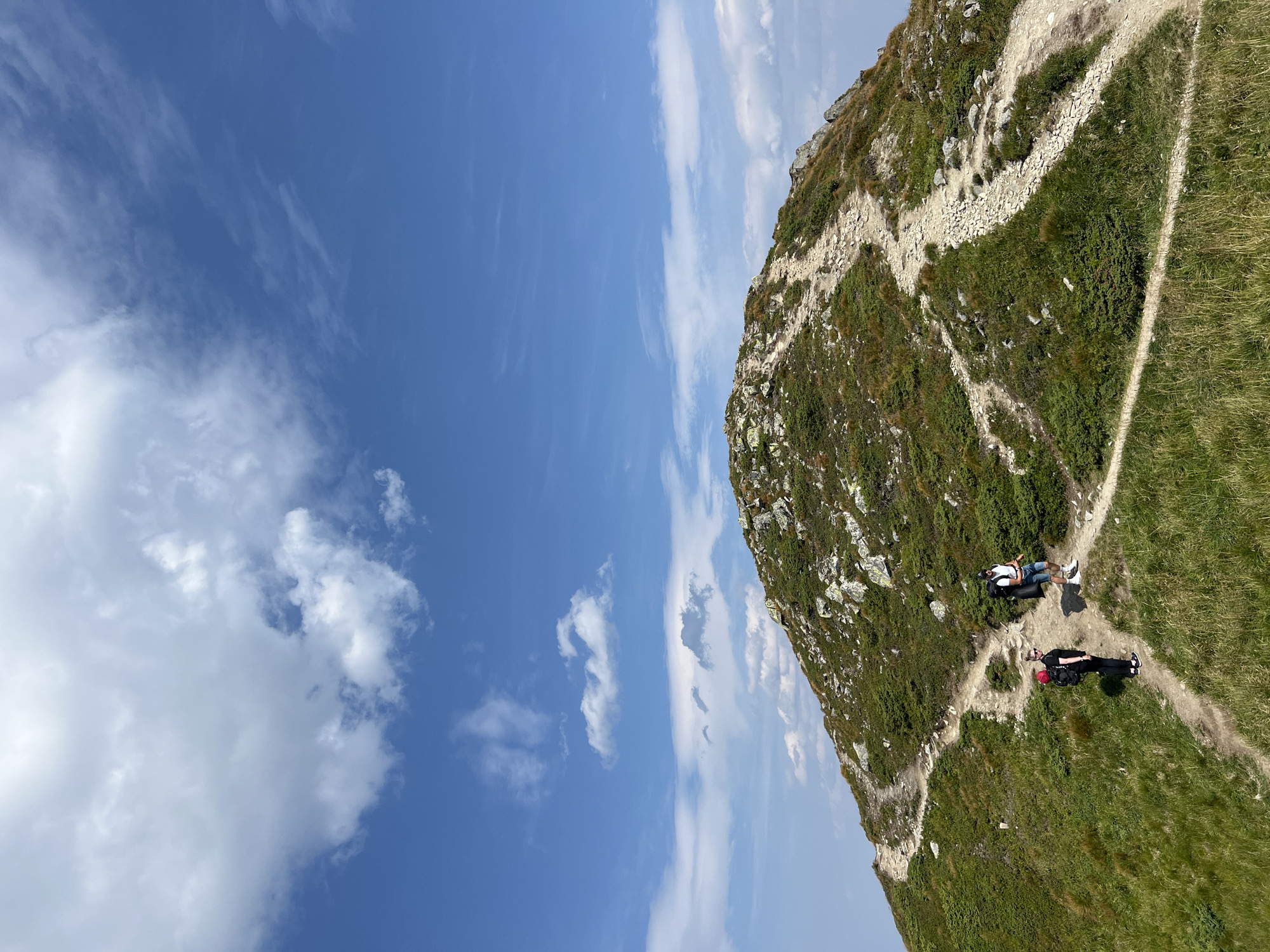 In the Carpathian Mountains near Vorokhta, Ukraine, two hikers ascend a rocky, grassy mountain path under a bright blue sky dotted with white clouds. The path is winding and appears well-trodden, leading towards the summit which is covered in rocks and low-lying green vegetation. One hiker, wearing a black shirt and black pants with a red backpack, pauses on the path, facing away from the camera. Another hiker, dressed in a white shirt and denim shorts with a large black backpack, walks further up the trail, his back to the camera. The overall scene depicts a clear, sunny day on a mountain hike, with the natural landscape dominating the frame.