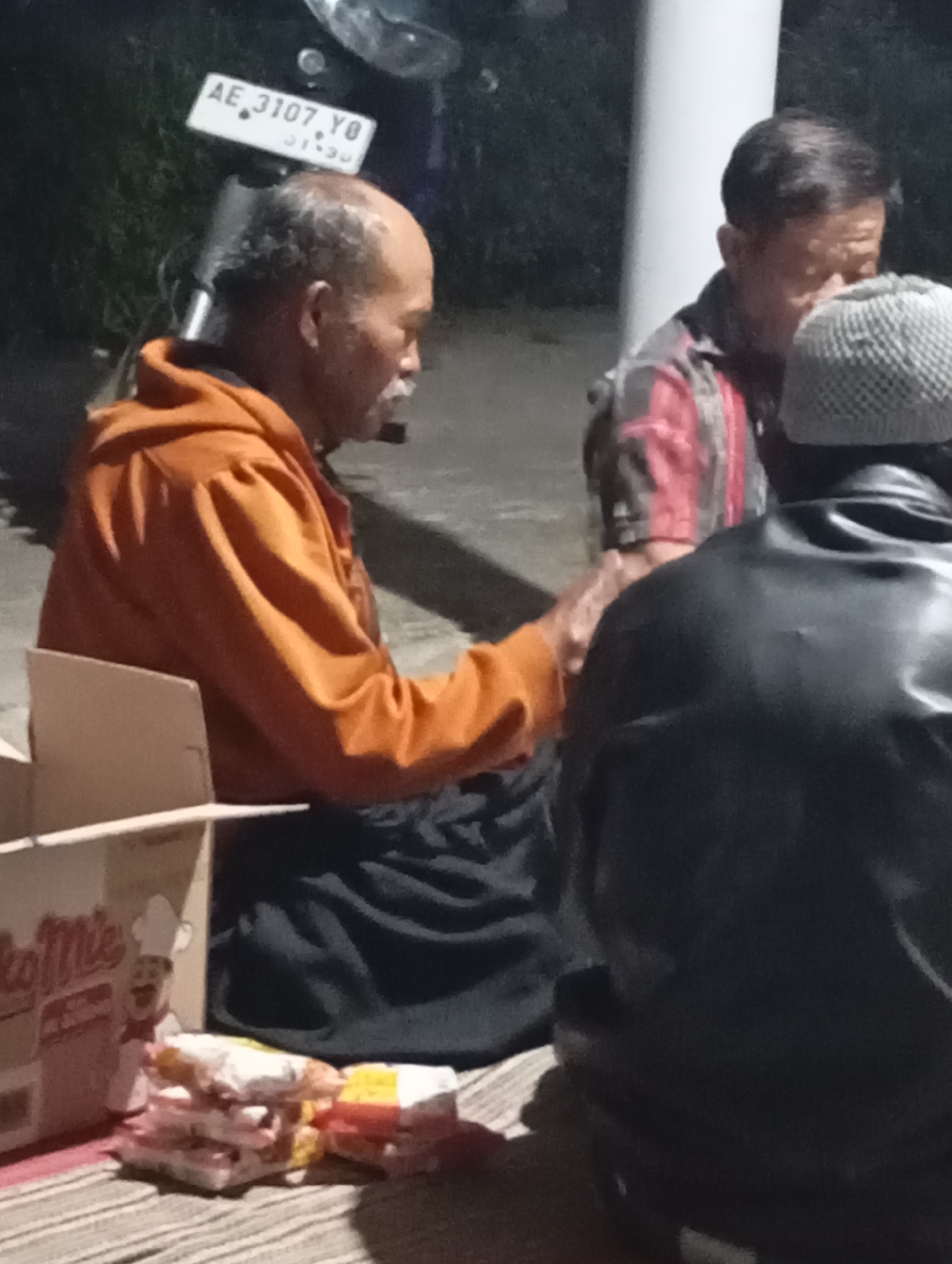 Three individuals are seated on the ground, possibly for a meal or gathering. The person on the left wears an orange hoodie and appears to be a bald, older man with a mustache, looking to his right. In the center, another man in a plaid shirt looks down, partially obscured by the person on the right. The person on the right wears a black leather jacket and a patterned beanie. In front of them are several packaged snacks and a cardboard box with a cartoon chef graphic on it, suggesting the presence of food items. A motorcycle with a license plate reading "AE 3107 YO" is visible in the background, and the overall setting appears to be an outdoor, dimly lit area. The presence of the license plate suggests this scene is likely located in Indonesia, as "AE" is a vehicle registration code for East Java, which includes the region of Wonogondo.