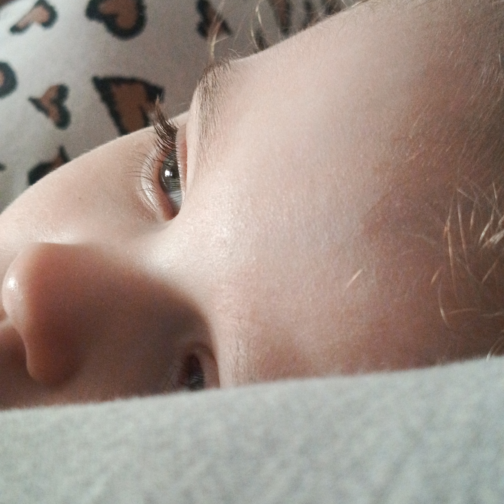 A close-up image shows the face of a child, specifically the nose, eye, and side of the face. The child appears to be lying down on a gray-colored surface, possibly a blanket or bedsheet. The background includes a patterned fabric with heart-shaped designs. There is no indication of location in the city of Марковский, Russia.