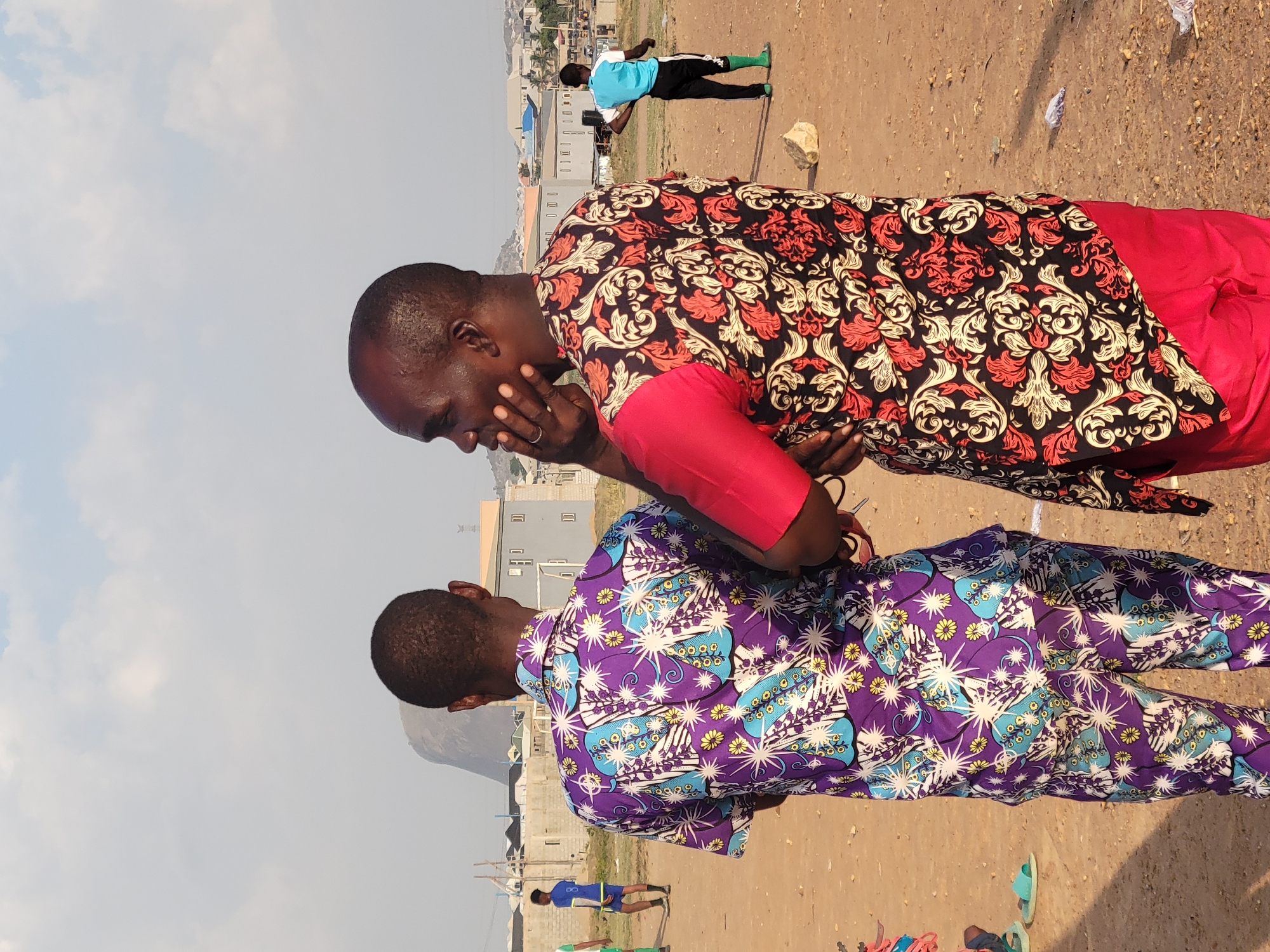 Two men are standing in a dirt clearing outside an urban area. One man is wearing a red-and-black patterned shirt with red sleeves and is touching his face. The other man is wearing a purple shirt with a light blue-and-white floral pattern. In the background are other people, buildings, and a hillside. The location is Ibo, Nigeria.