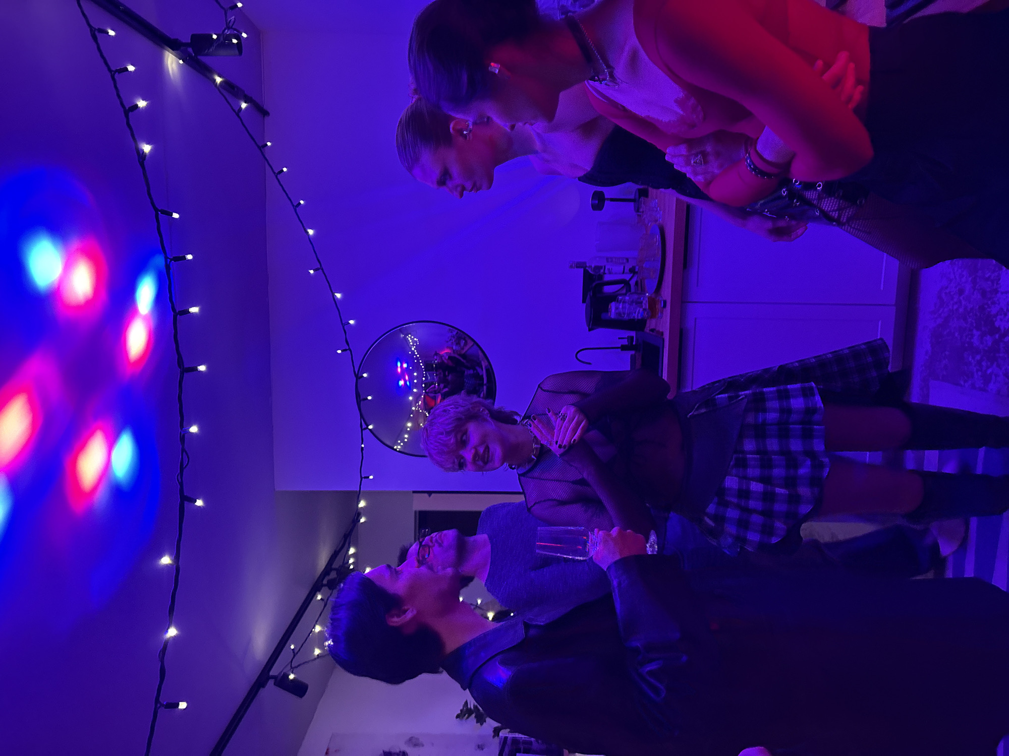 The photograph, illuminated by blue and red lights, shows a group of people in an indoor setting. Strings of small lights adorn the wall. In the foreground, a person in a black jacket holds a glass. Another individual, wearing a sheer top and plaid skirt, stands nearby. Further back, two women are visible near a counter area, seemingly engaged in conversation. The location is unknown.