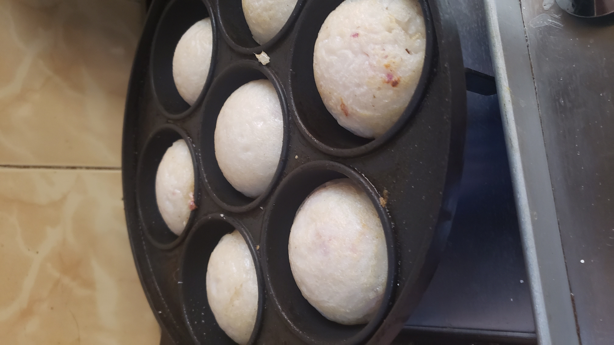 The image displays a close-up, slightly overhead view of a black, multi-mould cast-iron pan actively used for cooking. Seven round, light-colored food items, appearing to be mini-pancakes or spherical rice cakes (possibly "masa," a Nigerian delicacy), are visible nestled within the hemispherical depressions of the pan. These food items are mostly white or off-white, with some exhibiting faint reddish or brownish specks, suggesting the inclusion of other ingredients like onions or peppers, or slight browning from the cooking process. The pan itself shows some signs of use, with minor residue or discoloration.
To the left of the pan, a light brown or beige tiled surface is visible, possibly a kitchen counter or floor, featuring a subtle marbled pattern and dark grout lines. To the right, a darker, possibly metallic or plastic, grey-black surface suggests another kitchen appliance, likely a stove top, where the cooking is taking place.
The scene depicts an ongoing food preparation activity within a domestic kitchen setting, likely in Sabon Tasha, Nigeria. The time of day, weather, and any human presence or emotions are not discernible from the image. There is no visible text.