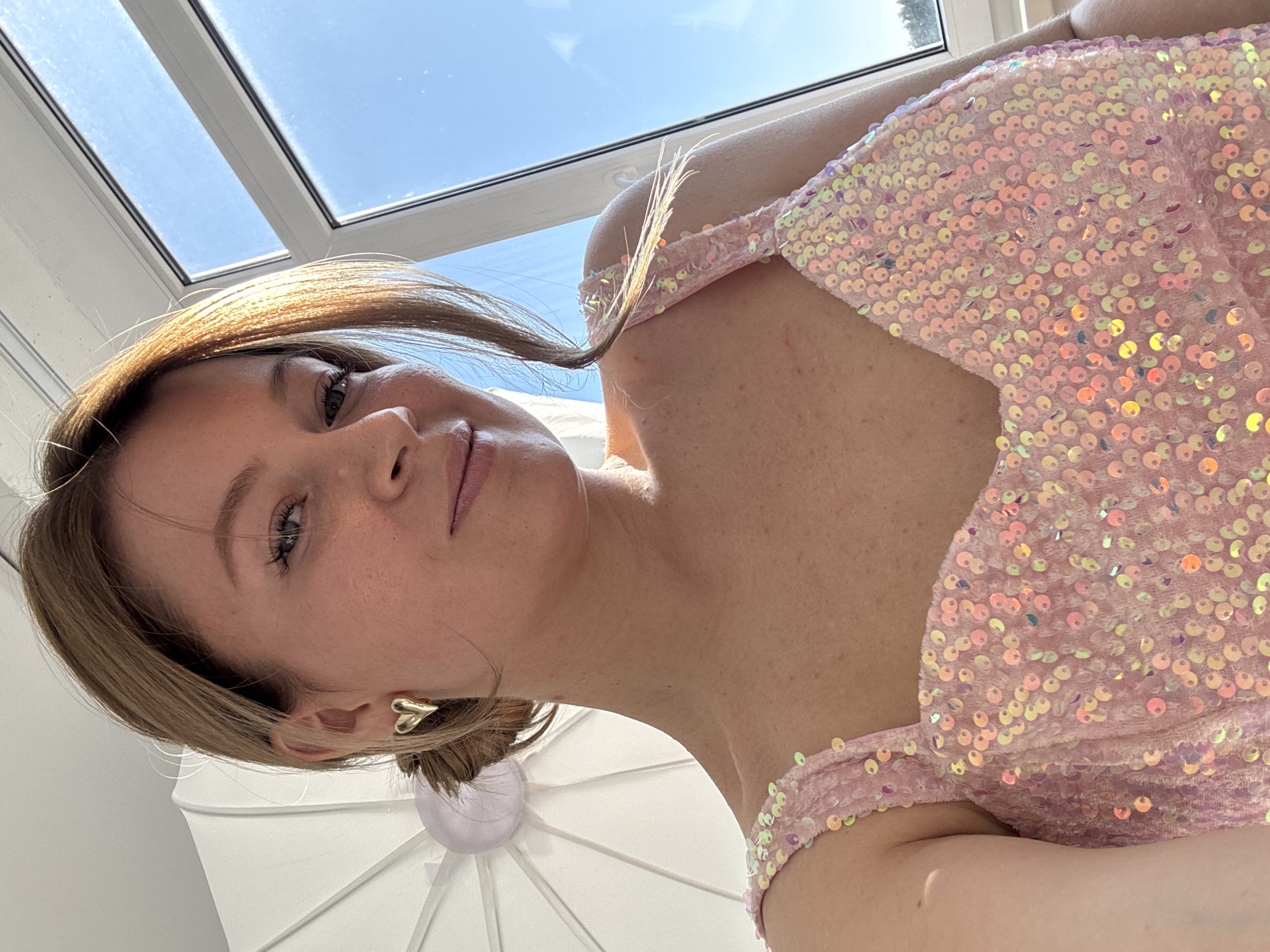 A woman poses indoors, possibly in a residential building. Her face is angled towards the camera. She has light brown hair, fair skin, and wears gold heart-shaped earrings. She is wearing a pink sequined top. Through a window above, a view of the sky is visible. There is a white object behind her, which appears to be an umbrella or reflector. The location is unspecified and there is no evidence to indicate it's in Chaikovsky, Russia.
