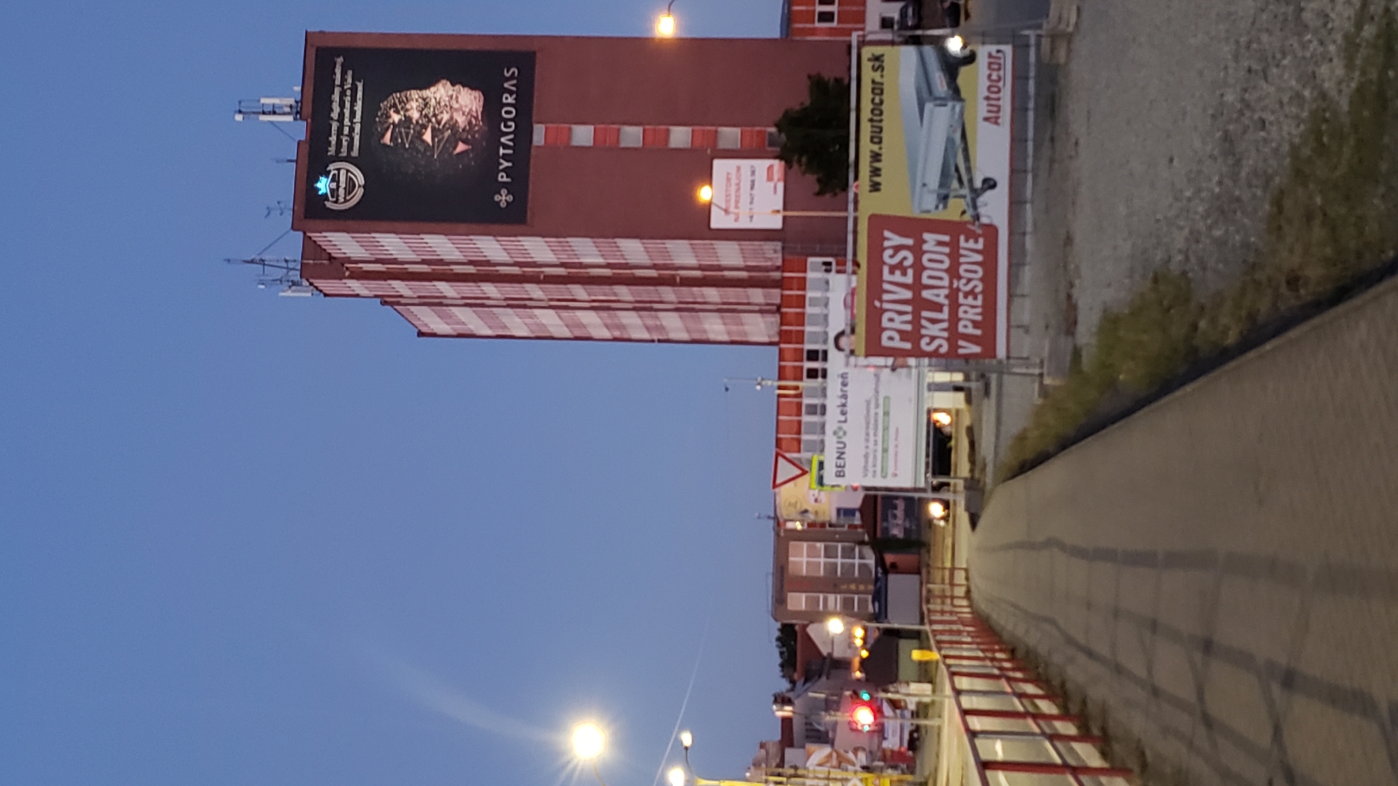 The image depicts a street scene in Prešov, Slovakia, near a tall, red and white striped building featuring a large advertisement for "PYTHAGORAS". The advertisement shows a stylized human head and text in Slovak. In the foreground, a pedestrian walkway with a red and white railing runs along the left side of the frame. Several billboards are visible, including one advertising "BENU Lekáreň" (BENU Pharmacy) and another for "PRÍVESY SKLADOM V PREŠOVE" (Trailers in stock in Prešov) with the website "www.autocar.sk". The sky is a clear blue, suggesting daytime. Traffic lights and streetlights are present, indicating an urban environment. Cars are parked or driving in the background. The overall scene captures a commercial area within the city of Prešov.