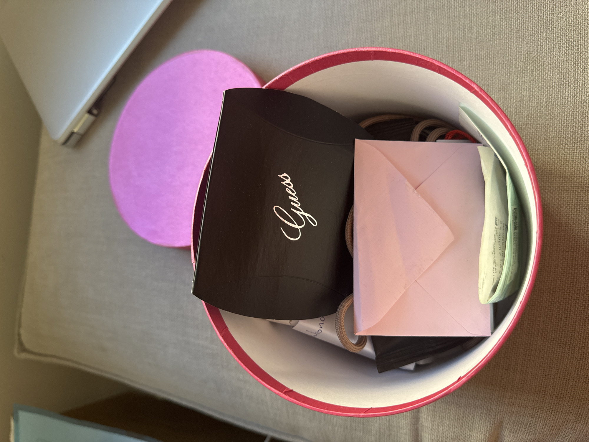 The image shows a top-down view of an open, round pink gift box. Inside the box are several items, including a black box with "Guess" printed on it in white cursive script, a small pink envelope, some hair ties, and what appears to be money or a gift card. The box is placed on a textured grey surface, possibly a couch or cushion. To the right of the box, a portion of a silver laptop is visible. In the background, a bright pink lid for the gift box is partially seen. There are no people visible in the image. The scene is indoors, with natural light illuminating the objects. The location is not specified to be in Murcia, Spain.