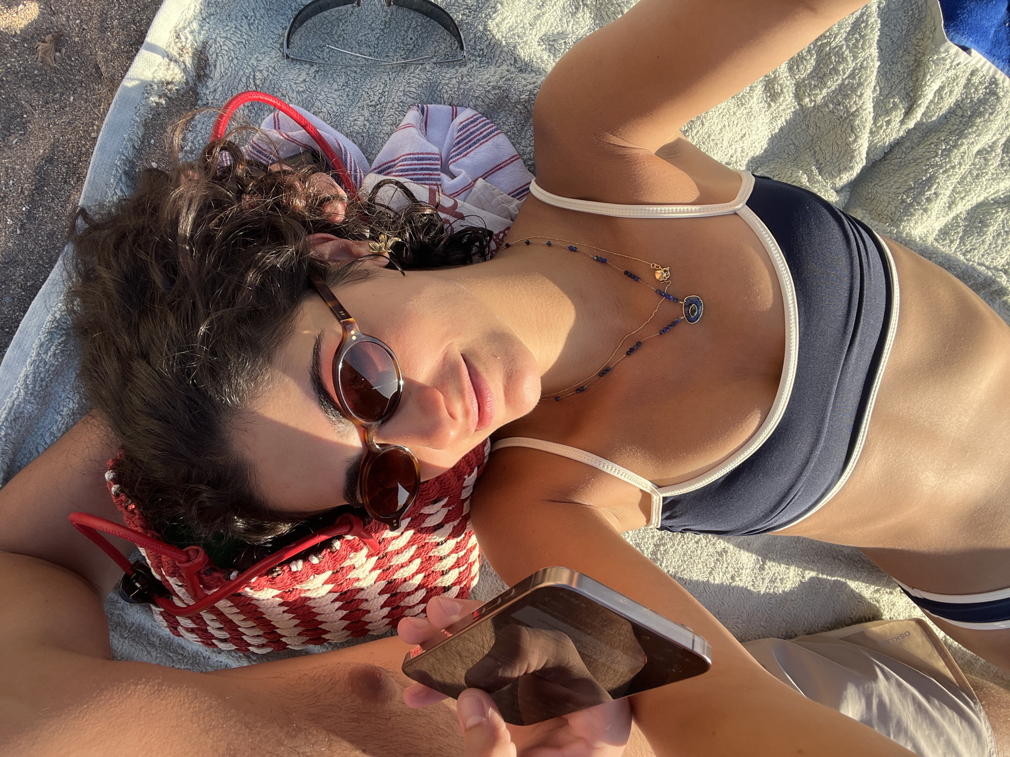 A woman with dark, curly hair and sunglasses lies on a light blue towel on a beach. She is wearing a navy blue bikini with white trim and a gold necklace with blue pendants. She is holding a silver smartphone in her left hand, with the screen facing away from the camera, reflecting her foot and the beach. To her left, another person's leg, possibly male, is visible. Next to her head is a red and white striped woven bag. A pair of sunglasses rests on the towel to her right. The background consists of dark sand, typical of a beach environment. The scene appears to be in the city of Sant Josep de sa Talaia, Spain.