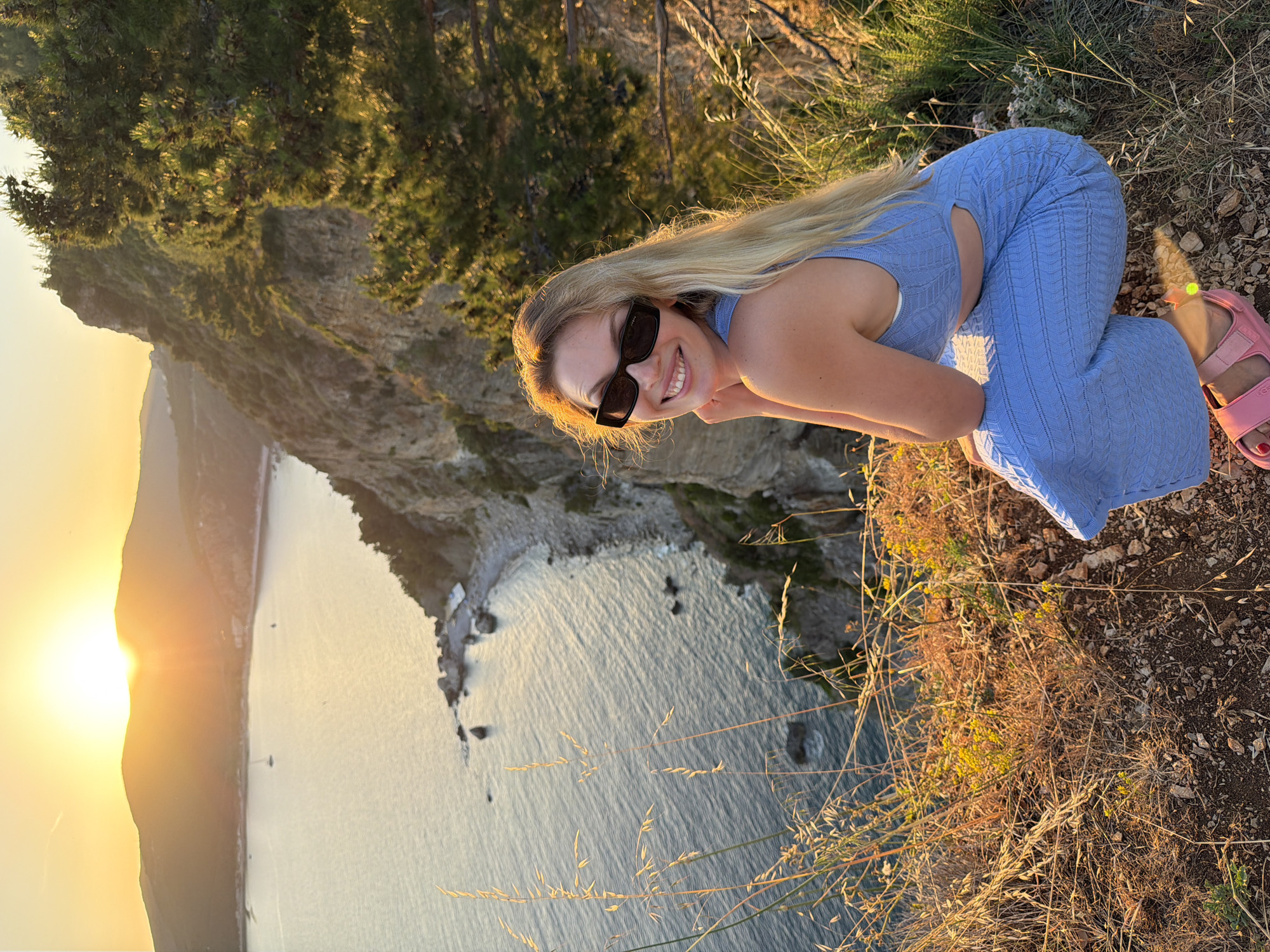 A young adult female is depicted crouching on a dry, rocky clifftop overlooking the Adriatic Sea at sunset. The subject is positioned in the lower right foreground, facing back and smiling towards the camera. She has long blonde hair and wears dark rectangular sunglasses. Her attire consists of a light blue two-piece knitted outfit, featuring a crop top and wide-leg trousers with cutouts at the waist. Pink Ugg-branded sandals are visible on her feet. Her left hand is raised, with fingers lightly touching her chin.
The immediate foreground consists of dry earth, small rocks, and sparse, straw-colored grasses and weeds, interspersed with small yellow flowering plants. The ground slopes sharply downwards towards a steep, rocky cliff face covered with green pine trees and shrubs.
In the midground, the sea below exhibits gentle ripples, reflecting the warm glow of the setting sun. A small, rocky beach or cove is visible at the base of the cliffs. Two sailboats are present on the water further out.
The background features a broad expanse of the sea and distant, dark mountains silhouetted against a vibrant orange and yellow sunset sky. The sun, a bright disk, is partially obscured by the mountain peaks on the horizon. The coastline forms a bay, characteristic of the Budva Riviera in Montenegro, with the view likely facing west from an elevated position. The scene is illuminated by golden hour light, creating a warm ambiance and backlighting the subject's hair.