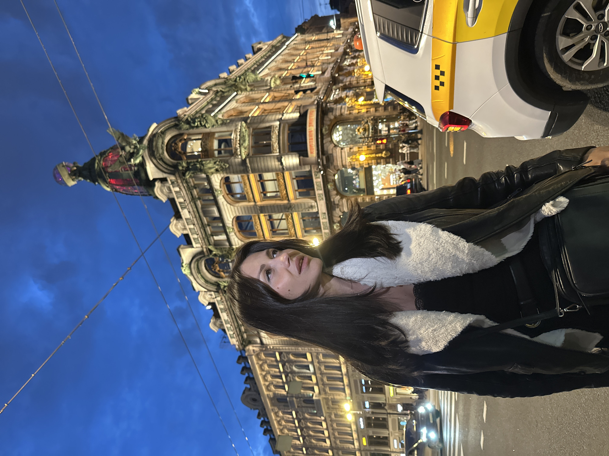 A woman stands on a city street at dusk in front of a historic building. The woman has long, dark hair and is wearing a black jacket with a white fur collar and a black top. She is looking to her right. To her right, a white taxi with yellow markings is parked. In the background, an ornate building with illuminated windows and a distinctive dome is visible. The sky is a deep blue, indicating twilight. The street appears to be a busy thoroughfare with other cars and pedestrians in the distance. The building in the background is likely the Singer House (Dom Knigi) on Nevsky Prospekt in Saint Petersburg, Russia, identifiable by its glass dome and architectural style.