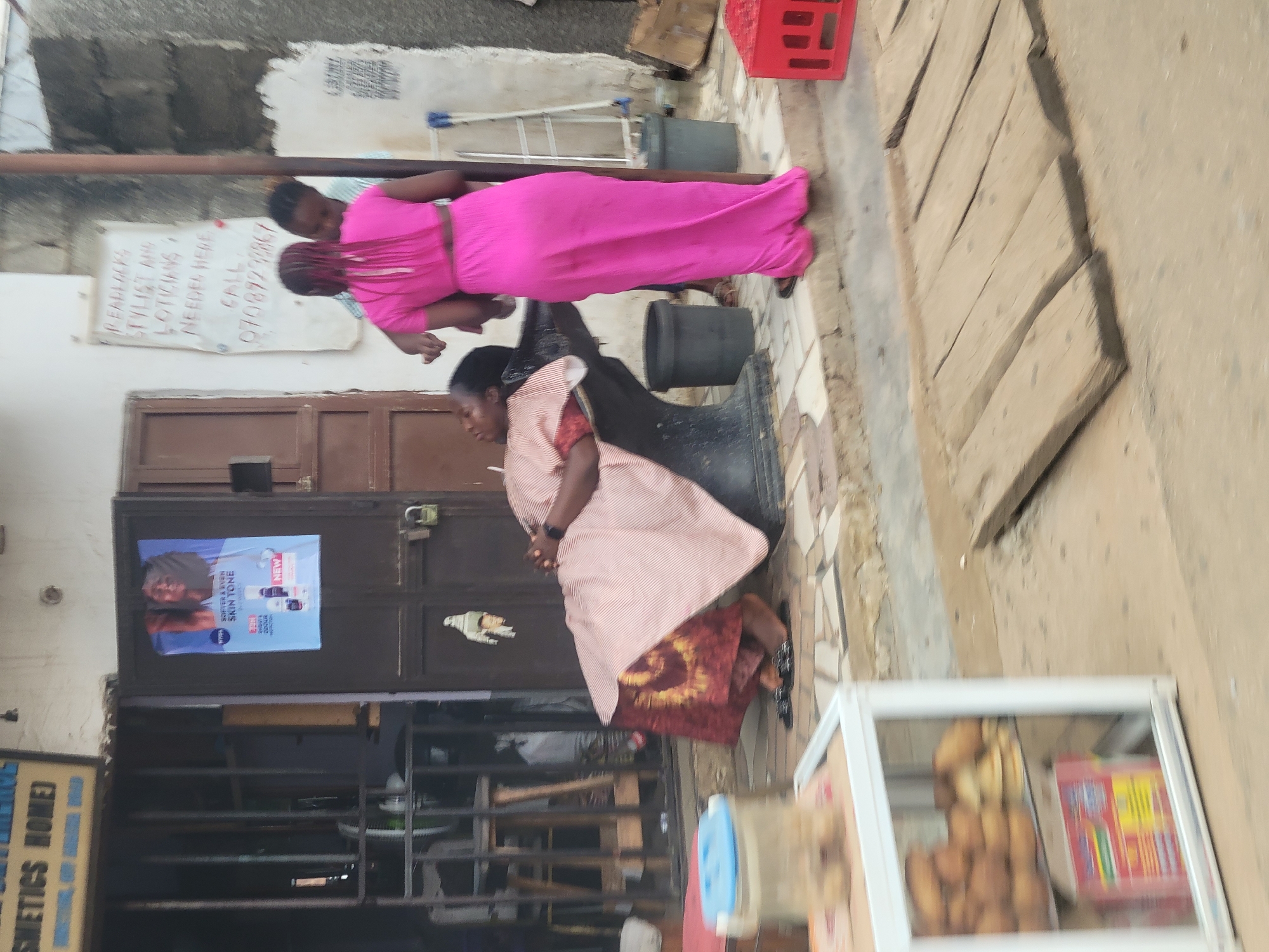 The image depicts a street scene in Katambe, Nigeria. In the foreground, a white display case filled with baked goods sits on the left. A wooden plank pathway is laid across the ground in the middle. Further back, a woman with a pink dress is styling the hair of another woman who is seated in a barber's chair, covered by a striped gown. Behind them, a building has a dark brown door with a poster on it. To the right of the door, a handwritten sign advertises "Readlocks Stylist and Lotions Needed Here. Call 07089229967." Several dark gray trash bins are visible, along with a red plastic crate. The overall environment appears to be an outdoor commercial area.
