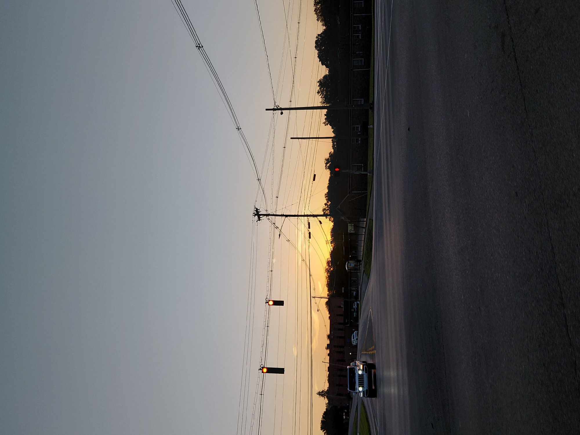 The image depicts a street scene at dusk. The sky is a pale gray, transitioning to a warm orange and yellow hue near the horizon, indicating the sun is setting. Silhouetted against the colorful sky are trees and buildings, with power lines crisscrossing the frame. Two traffic lights, both displaying a red signal, are visible. A white SUV is driving on the asphalt road, its headlights illuminated. The road appears to be a multi-lane street with painted lane dividers. The setting suggests an urban or suburban environment in the United States. The overall mood is peaceful, with the fading light creating a serene atmosphere. No visible text is present in the image.