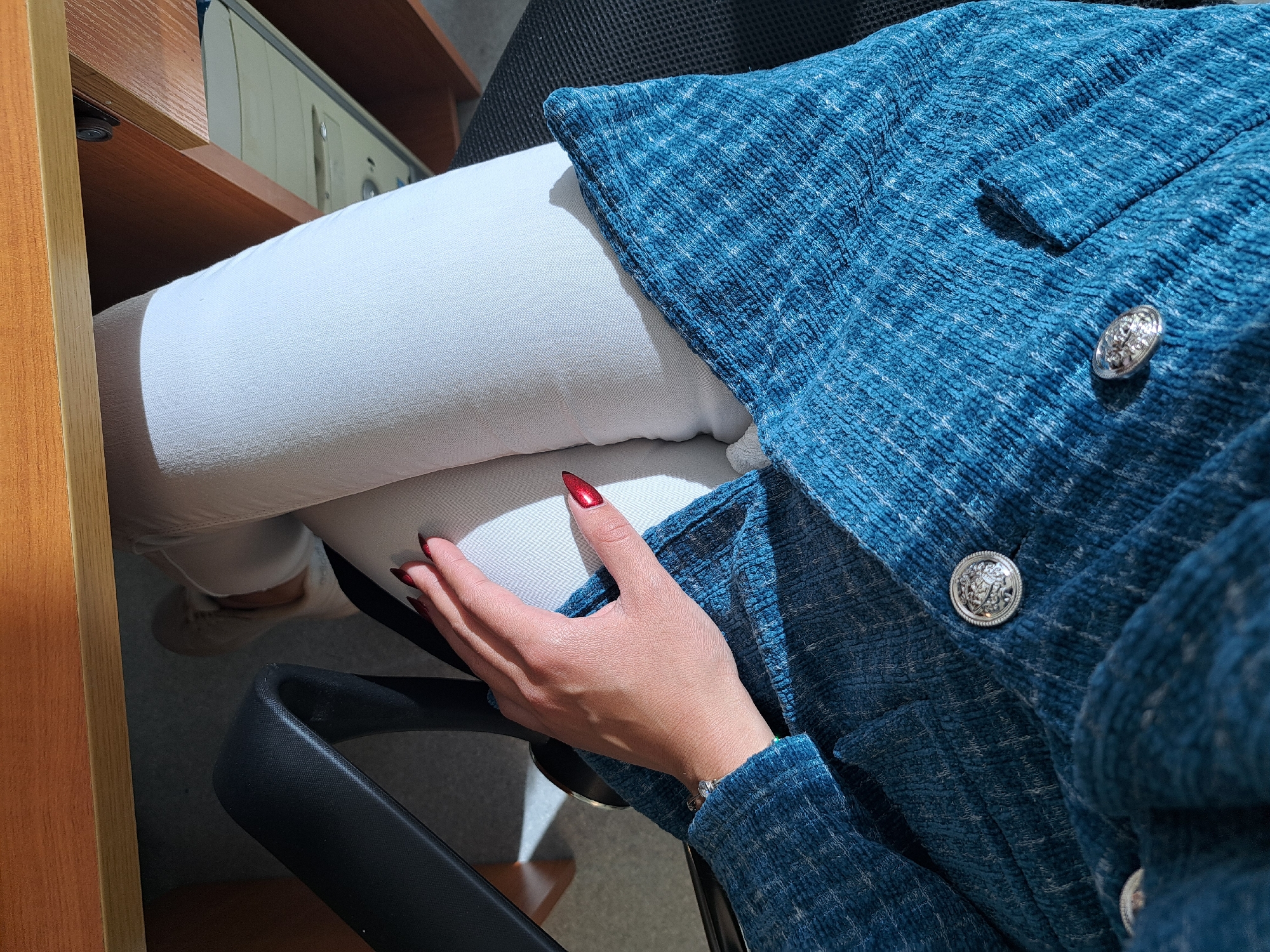 The image shows a person sitting in an office chair, wearing white pants and a blue tweed jacket with ornate silver buttons. The person has red nail polish and is wearing a bracelet. A wooden desk and computer are visible in the background. The location is likely indoors, possibly an office in Sofia, Bulgaria.