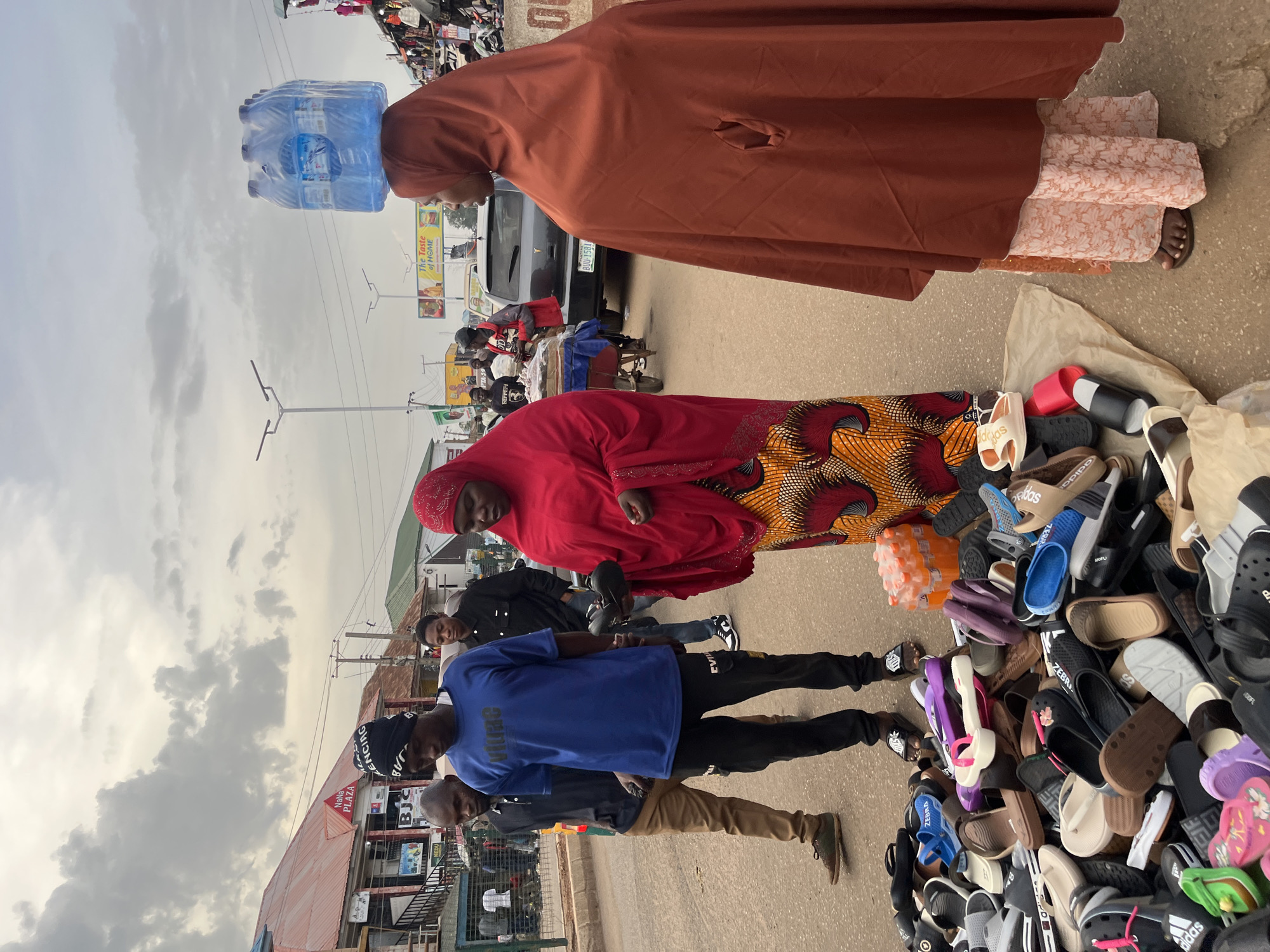 In Bukuru, Nigeria, an outdoor market scene unfolds under a cloudy sky. A woman wearing a burnt orange hijab and a patterned skirt balances a pack of bottled water on her head, walking alongside a woman in a vibrant red hijab and a patterned dress who is interacting with a vendor. The vendor, a man in a blue t-shirt and black trousers, appears to be showing items to the woman in red. Behind them, other individuals are seen in the bustling street, with cars and market stalls visible in the background. A large collection of assorted footwear, including sandals and flip-flops, is displayed on the ground in the foreground, suggesting a shoe vendor's stall. The overall environment suggests a typical day in a busy Nigerian marketplace.