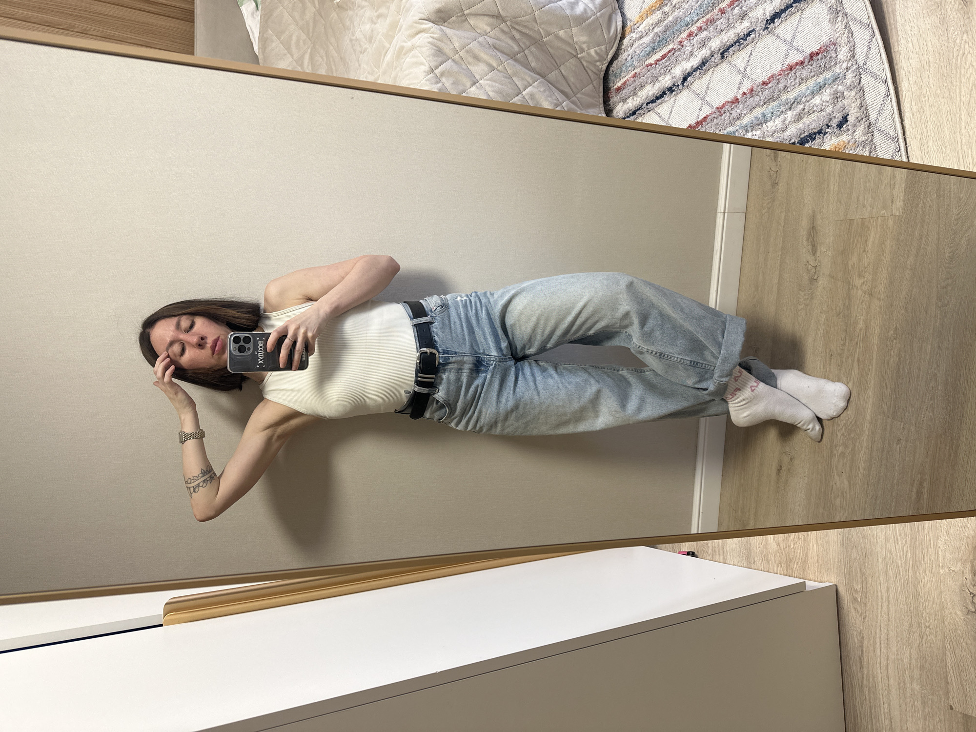 A woman is visible in a mirror. She is wearing a white tank top and light-wash blue jeans with a black belt. She is lying down, and holding a phone in her right hand, near her face, while resting her left hand on her forehead. She has short, dark hair and is wearing a watch on her left wrist. A tattoo is visible on her left forearm.
The reflection shows a portion of a bed with a quilted white cover. A patterned rug is visible above the bed. A white cabinet is positioned below the mirror. The floor is made of light-colored wood.
The image was not taken in Votkinsk, Russia.