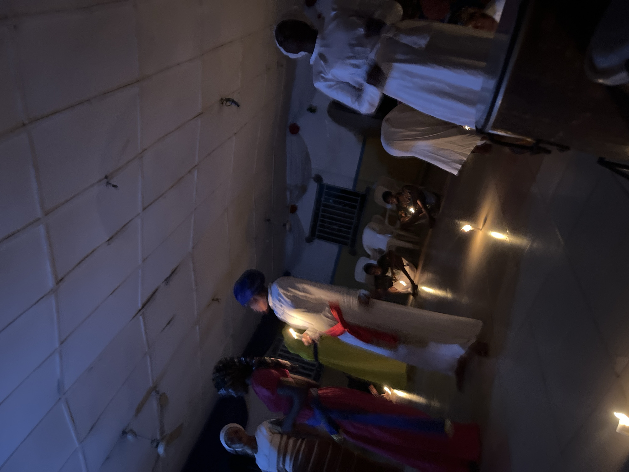The image depicts an indoor gathering, illuminated primarily by candlelight, at an unknown location within an unspecified city. The scene is characterized by dim, ambient lighting, with a blueish cast from unseen overhead sources complementing the warm glow of numerous lit candles.
The environment features a ceiling composed of light-colored square panels, from which a three-bladed ceiling fan is suspended in the upper left. The walls are light-colored, possibly blue and white, with a rectangular window fitted with security bars visible on the left side. Red circular ornaments and white fabric swags adorn the far wall near the ceiling. The floor is a dark, smooth, and reflective surface, showing distinct reflections of the candle flames. Several white plastic or metal chairs are scattered throughout the space, some unoccupied, some partially obscured. A large, brown, cylindrical object, resembling a drum, is partially visible in the bottom right corner.
Approximately seven to ten individuals are present, most appearing to be adults and dressed in long, loose-fitting garments, some with head coverings. In the left-midground, a person wearing a white top and a light head covering stands facing right, partially obscured. Beside them, a person in a long pink or red dress with a blue sash and braided hair stands, also facing right. To their right, a person in a long yellow-green dress is partially visible, holding a lit candle. Adjacent to them, a person in a long white robe with a red sash and a blue head covering holds a lit candle, facing left. On the right side of the frame, a person in a long white robe and a white head covering stands facing left, with their hands slightly raised. Another person in similar white attire is partially visible on the far right. Two individuals are seen crouching or sitting on the floor, each holding a lit candle; one is near the window, wearing a patterned shirt, and another is closer to the center, wearing a dark top. The individuals are positioned among a scattering of lit white pillar candles placed directly on the floor. Most standing individuals appear to be engaged in a shared activity or conversation.