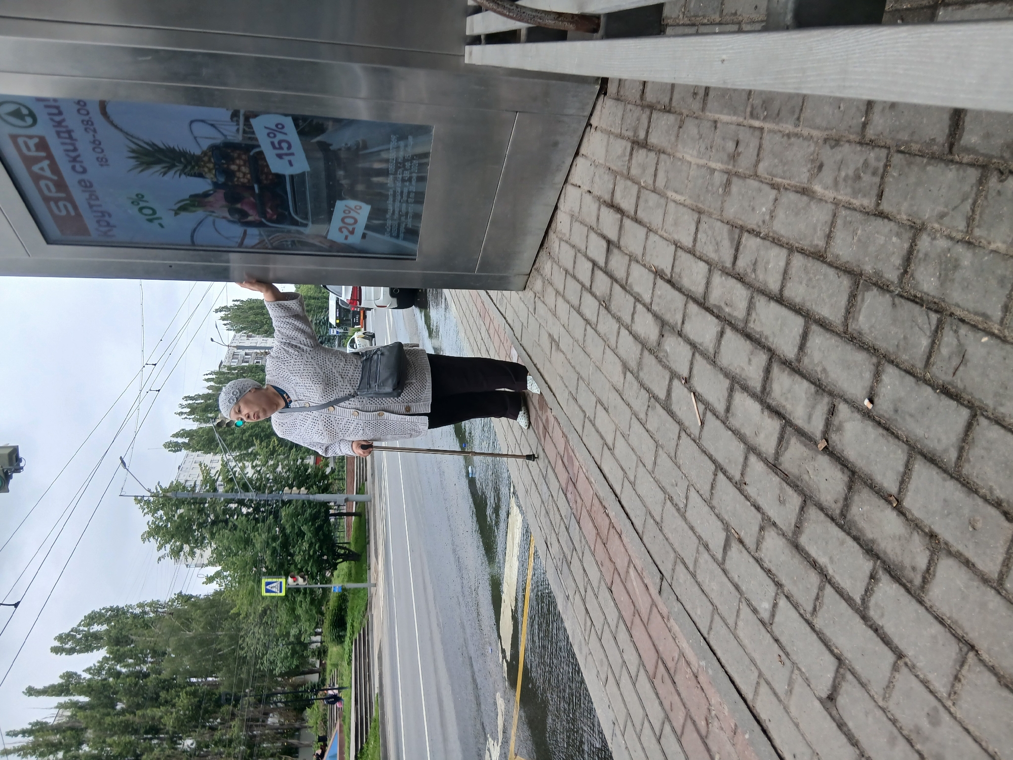 The image depicts an elderly woman standing on a sidewalk next to a large advertising display. The woman is wearing a light-colored, patterned sweater, dark pants, and a matching headscarf. She is holding a cane in her right hand and her left hand is resting on the advertising display. The advertising display is for SPAR and shows a sale with percentages off. Behind the woman, a street with traffic is visible, as well as trees and buildings in the background. The sidewalk is paved with bricks. The scene appears to be on a cloudy day. The location is identified as Nizhny Novgorod, Russia, based on the SPAR advertisement which is a Russian supermarket chain. The image is taken from a low angle, looking up at the woman and the display.
