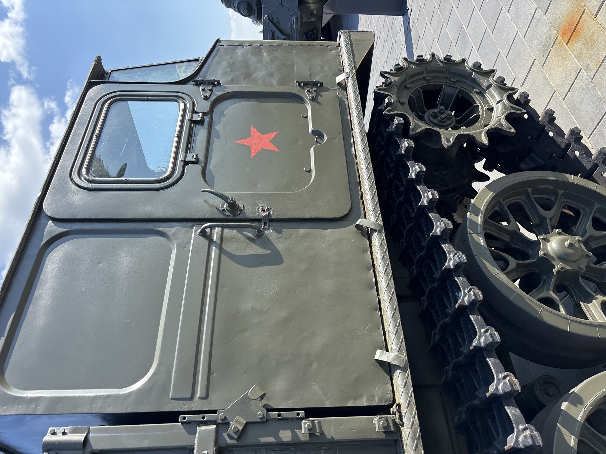 The image shows a side view of a green, tracked military vehicle. The vehicle is olive drab and has a prominent red star emblem on its side. It is parked on a paved surface, likely an outdoor exhibition or museum. In the background, another military vehicle, possibly a tank, is partially visible. The sky is blue with some clouds. The vehicle appears to be a tractor or transport vehicle. The scene is set at the Ozyaritska-Slabadski rural council in Belarus.