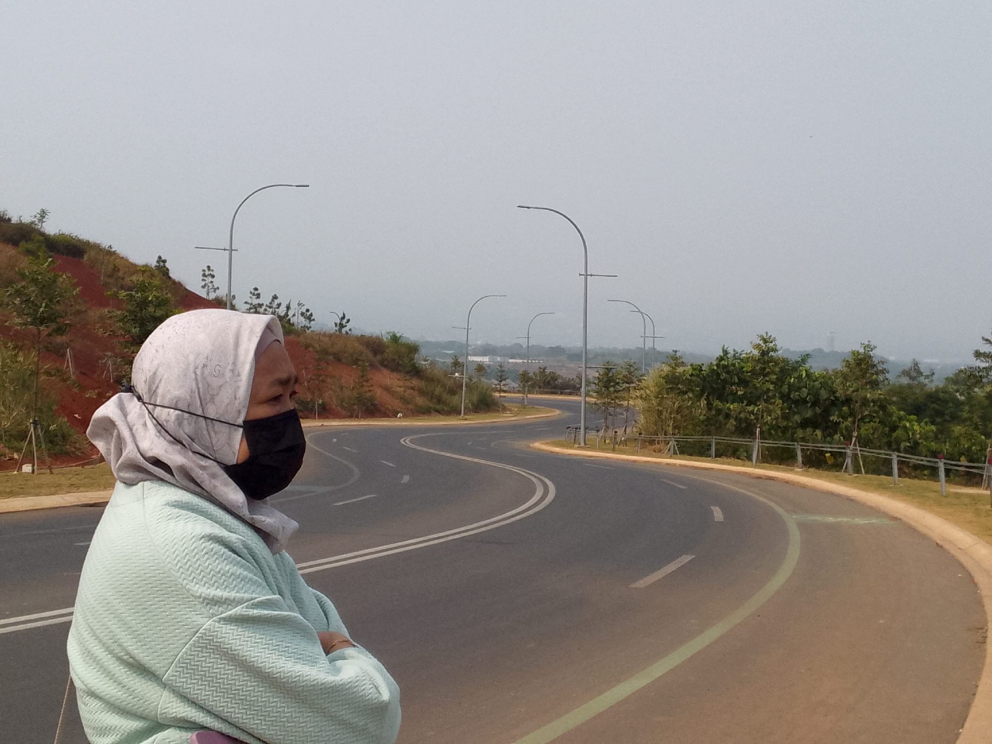 A woman wearing a light blue hijab and a black face mask is standing on the side of a winding asphalt road. She is looking to her right, with her arms crossed. The road has white lane markings and a thin green line along its edge. The surroundings are hilly with sparse vegetation, including trees and bushes. Several modern streetlights with curved poles are visible along the road. In the background, the sky is overcast and hazy, obscuring distant details. The location is identified as Bojonghaleuang, Indonesia.