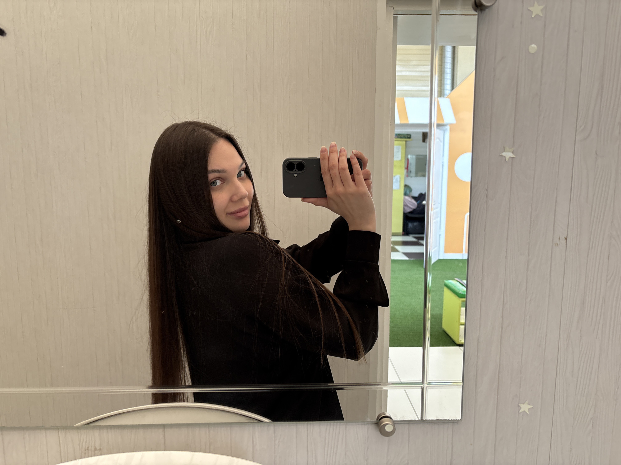 The image shows a woman with long, dark brown hair, fair skin, and blue eyes taking a mirror selfie. She's holding a dark-colored smartphone with a dual-lens camera, partially obscuring her face. She is wearing a dark-colored blouse or jacket. The setting appears to be a bathroom or similar space with light beige walls and a wooden door or panel to the right, adorned with small star-shaped decorations. Reflected in the mirror is a view into what appears to be a children's playroom with a playhouse structure in orange and white, a green carpet, and a lime-green bench or toy storage unit. The location is likely Chaikovsky, Russia.