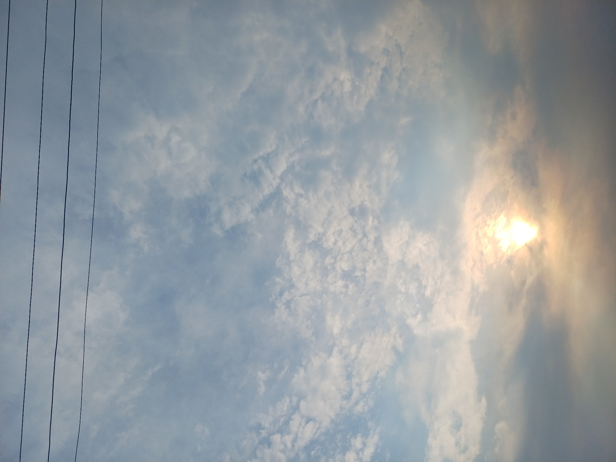 The image shows a sky with clouds and the sun. There are also power lines visible in the upper part of the image. The sun is partially obscured by clouds, creating a hazy glow. The clouds are varied in texture, with some appearing wispy and others more dense. The overall lighting suggests it might be late afternoon or early morning. There are no people or any other identifiable objects or structures within the frame. The location in Trypillia, Ukraine is not evident from the visual information provided in the image.