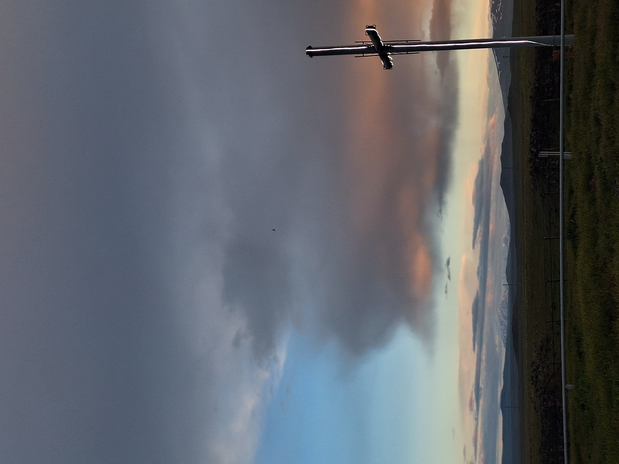 The image shows a landscape under a dramatic sky. Dark storm clouds dominate the left, contrasting with lighter, colorful clouds on the right near the horizon, possibly indicating sunset or sunrise. A metal structure resembling a cross is positioned near the top center of the frame. The landscape features green fields or vegetation, possibly a hillside, and what appears to be a mountain range in the distance. A fence line runs along the edge of the field. There is a bird in the sky. The location in the city of Anipemza, Armenia is not evident in the photo.